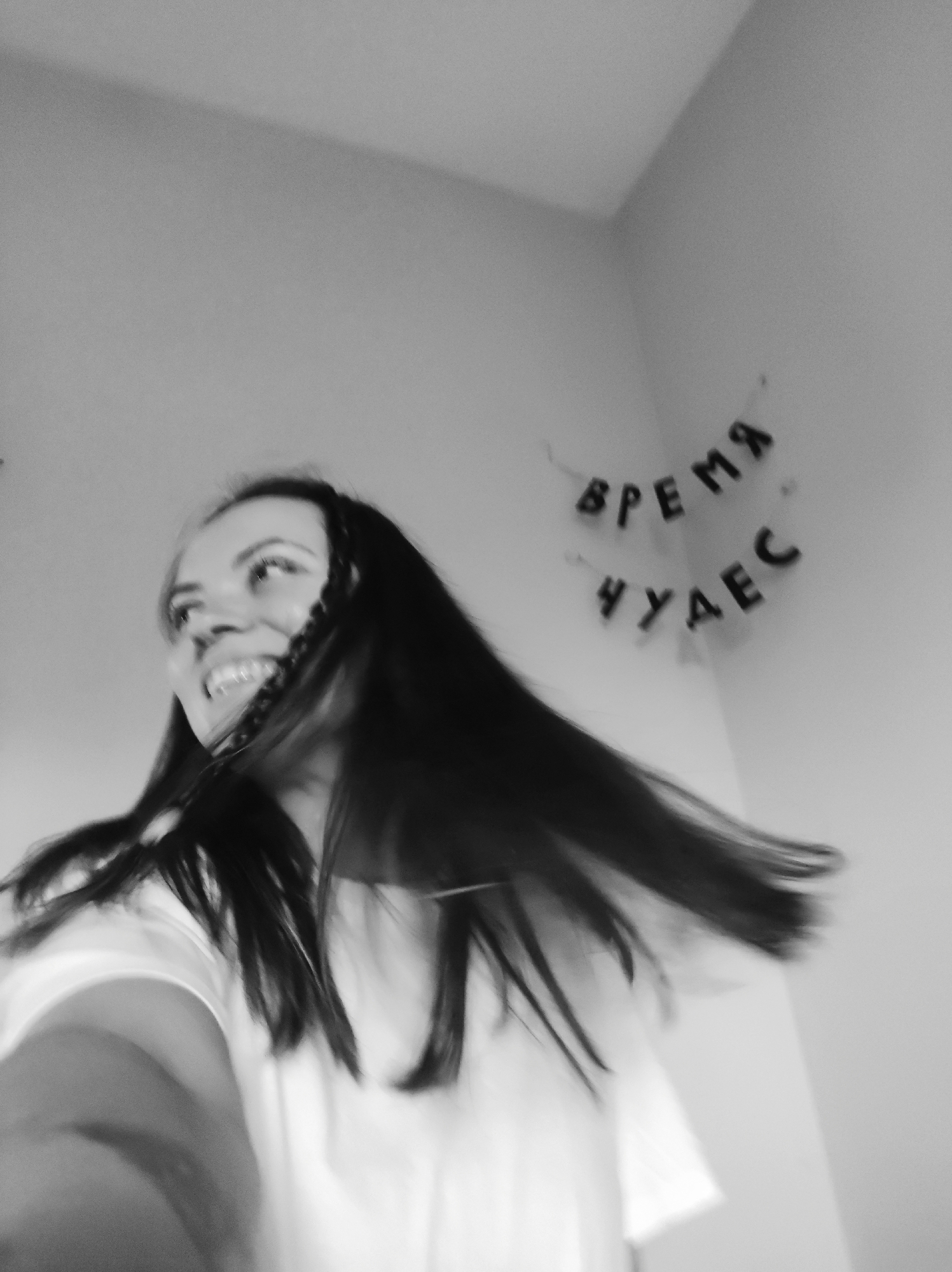 A monochromatic, motion-blurred photograph depicts a young adult female captured in a dynamic pose within an indoor room. The woman, positioned in the lower-left, has long, dark hair that appears to be in motion, flowing towards the right. Her face is in profile, gazing upward and to the right with a wide, open-mouthed smile, revealing teeth. She is wearing a light-colored garment, likely a t-shirt. The background is a plain, light-colored wall with a visible corner. Hung on the wall in the upper-right quadrant are two strings of dark letters. The upper string spells "ВРЕМЯ" (VREMYA) and the lower string spells "ЧУДЕС" (CHUDES), translating from Russian to "TIME OF MIRACLES" or "TIME FOR WONDERS." The image conveys a sense of exhilaration and joy, amplified by the significant motion blur affecting the subject and suggesting rapid movement. The location is an interior space in Unknown, Unknown.