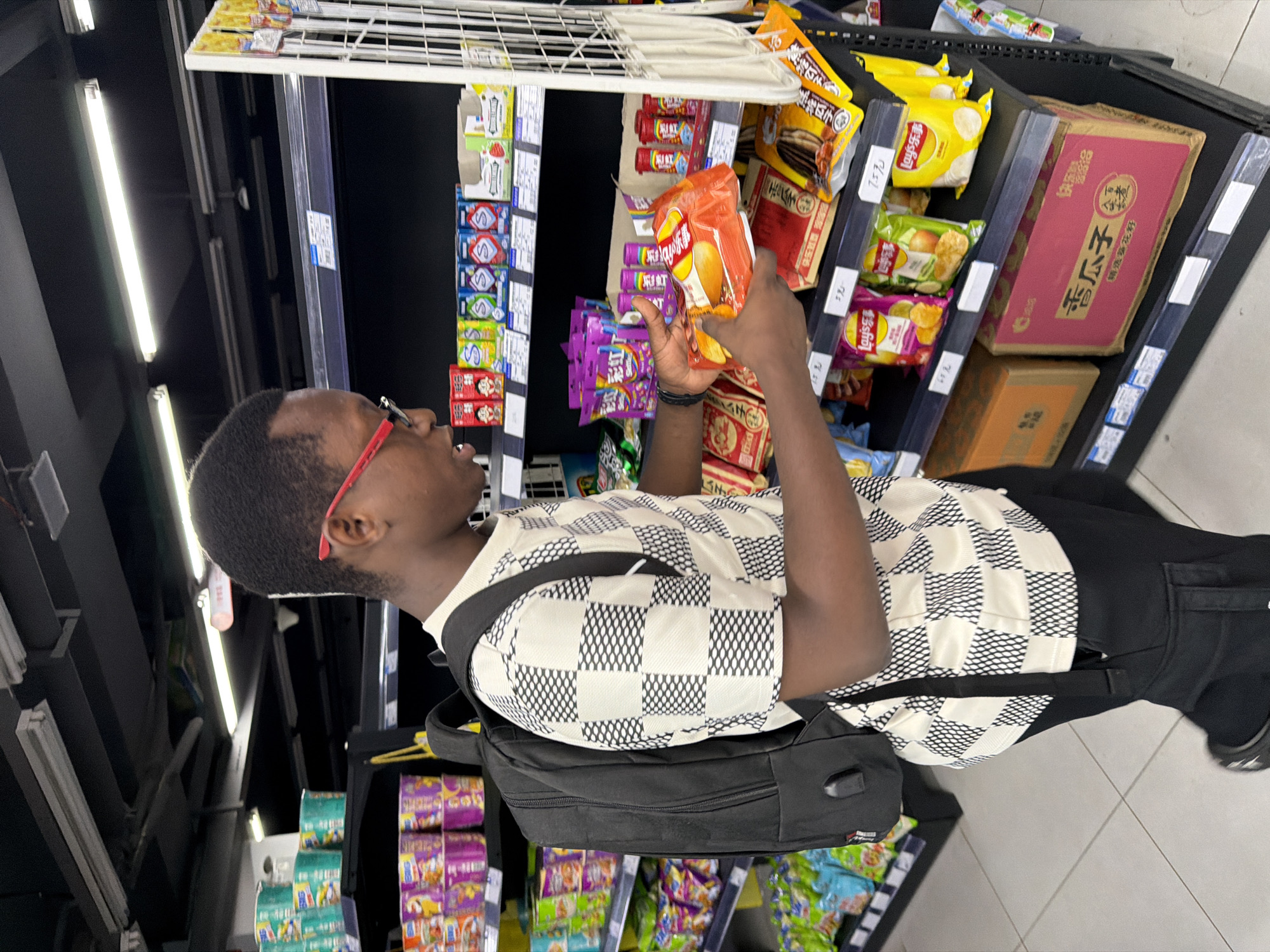 A young Black male wearing a black backpack and a black and white checkered t-shirt stands in an aisle of a brightly lit supermarket, holding a bag of chips. He is wearing red-rimmed glasses and is looking down at the chips in his hands, which are orange and yellow. The shelves behind him are stocked with various snack items, including colorful bags of chips and other packaged goods. The floor is tiled and appears to be clean. The overall environment suggests a typical retail setting.