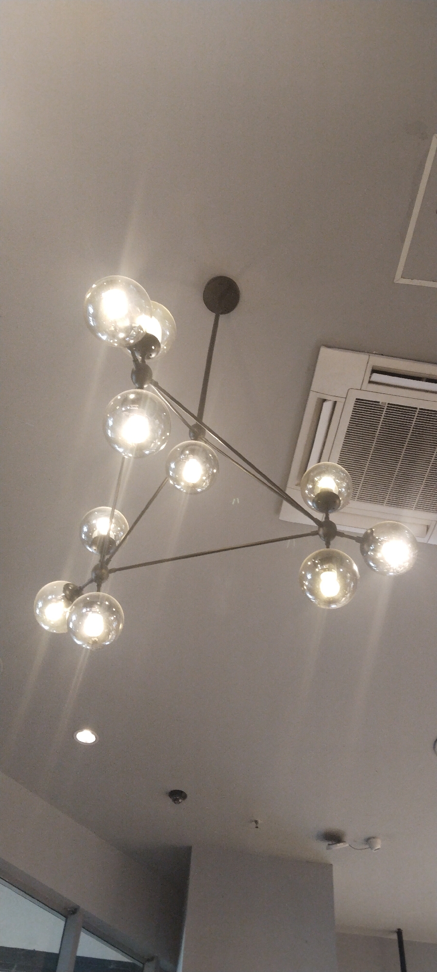 This image captures a modern chandelier with multiple glass bulb fixtures suspended from a grey ceiling. The chandelier has a geometric design with thin metal rods branching out to hold the bulbs. Some of the bulbs are illuminated, casting a warm glow. In the background, a white air conditioning unit is visible, also mounted on the ceiling. The overall environment appears to be an indoor space, possibly a commercial establishment or a well-lit room. The specific location in Semarang, Indonesia, cannot be determined from the visual information provided in the image alone.