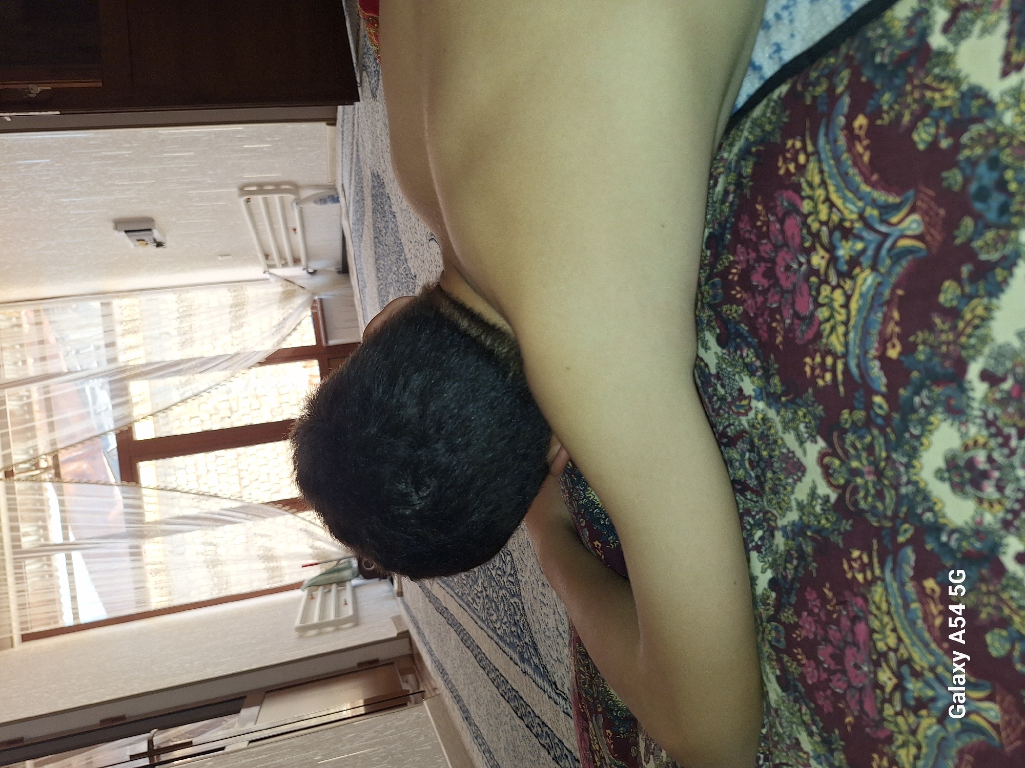 In a room in Charkhin, Uzbekistan, a shirtless person lies face down on a patterned rug. The person's dark hair is visible, and their arm is extended. The room features a large window with sheer curtains, a doorway with a dark wooden door, and a patterned carpet. The overall lighting suggests it is daytime.