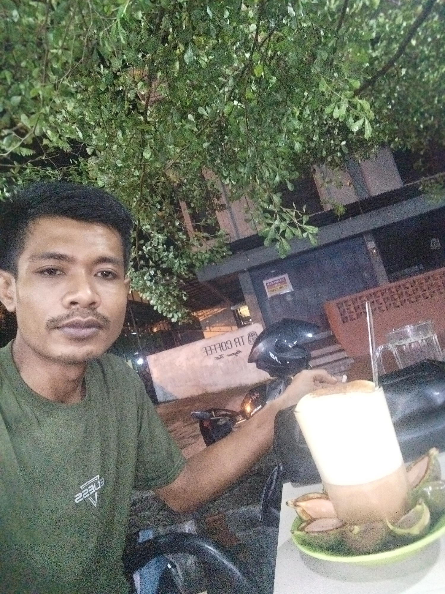 A man is taking a selfie outdoors in the city of Lhokseumawe, Indonesia. The man is wearing an olive green t-shirt. He is sitting with a motorcycle in front of him. Resting on the motorcycle is a drink, possibly coffee, in a tall glass surrounded by sliced avocado in a green bowl. In the background is a building with signage in reverse reading “TB Coffee”. Foliage is visible above. The lighting suggests it is nighttime.