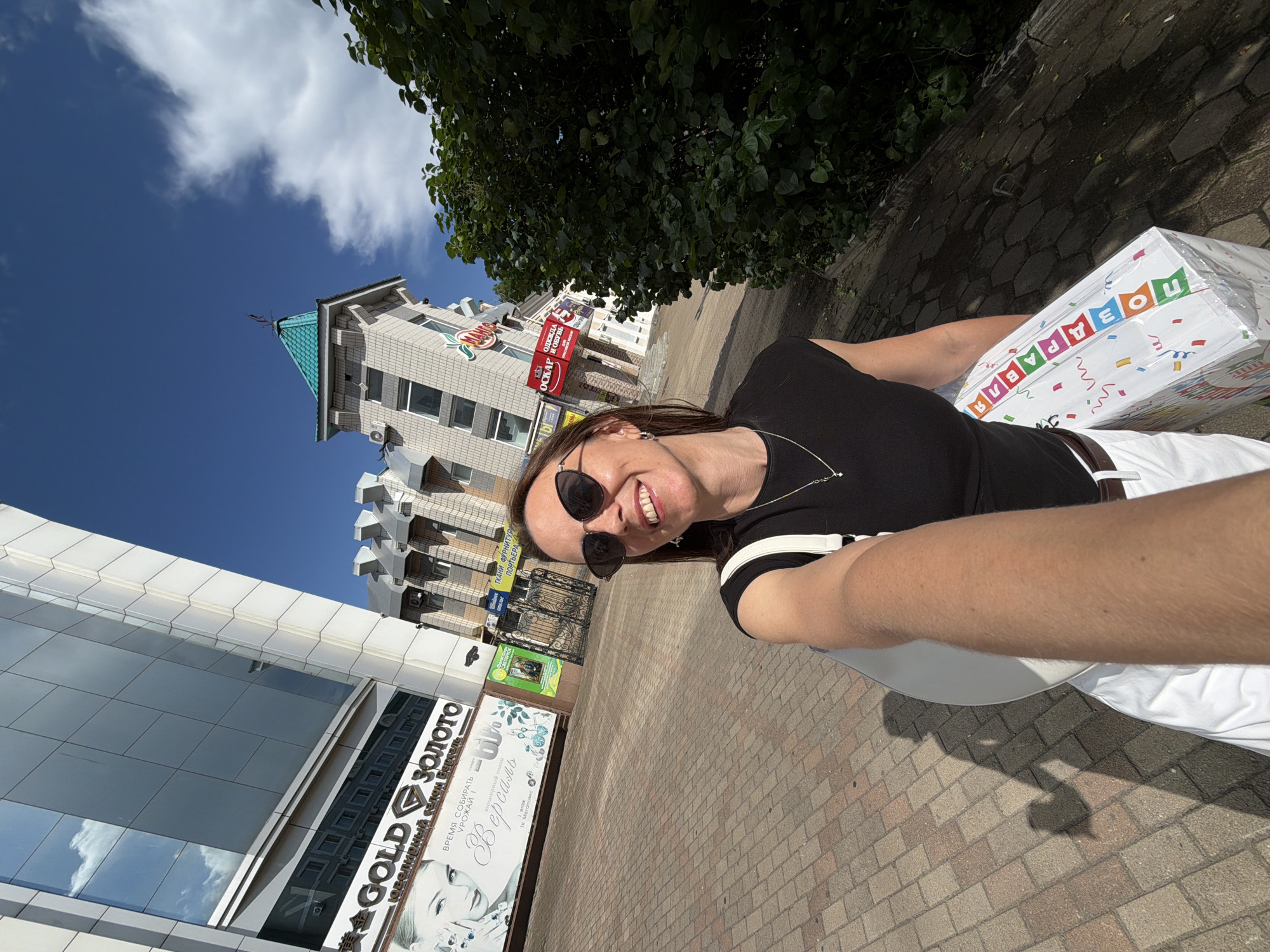 A woman is taking a selfie outdoors in front of buildings in Birobidzhan, Russia. The woman is smiling and wearing sunglasses, a black top, and white pants. She is holding a white bag and a large, colorful gift box. In the background, there are modern and older-style buildings, including a building with a green roof. The sky is blue with some clouds. The ground is paved with bricks. The image is taken on a sunny day.