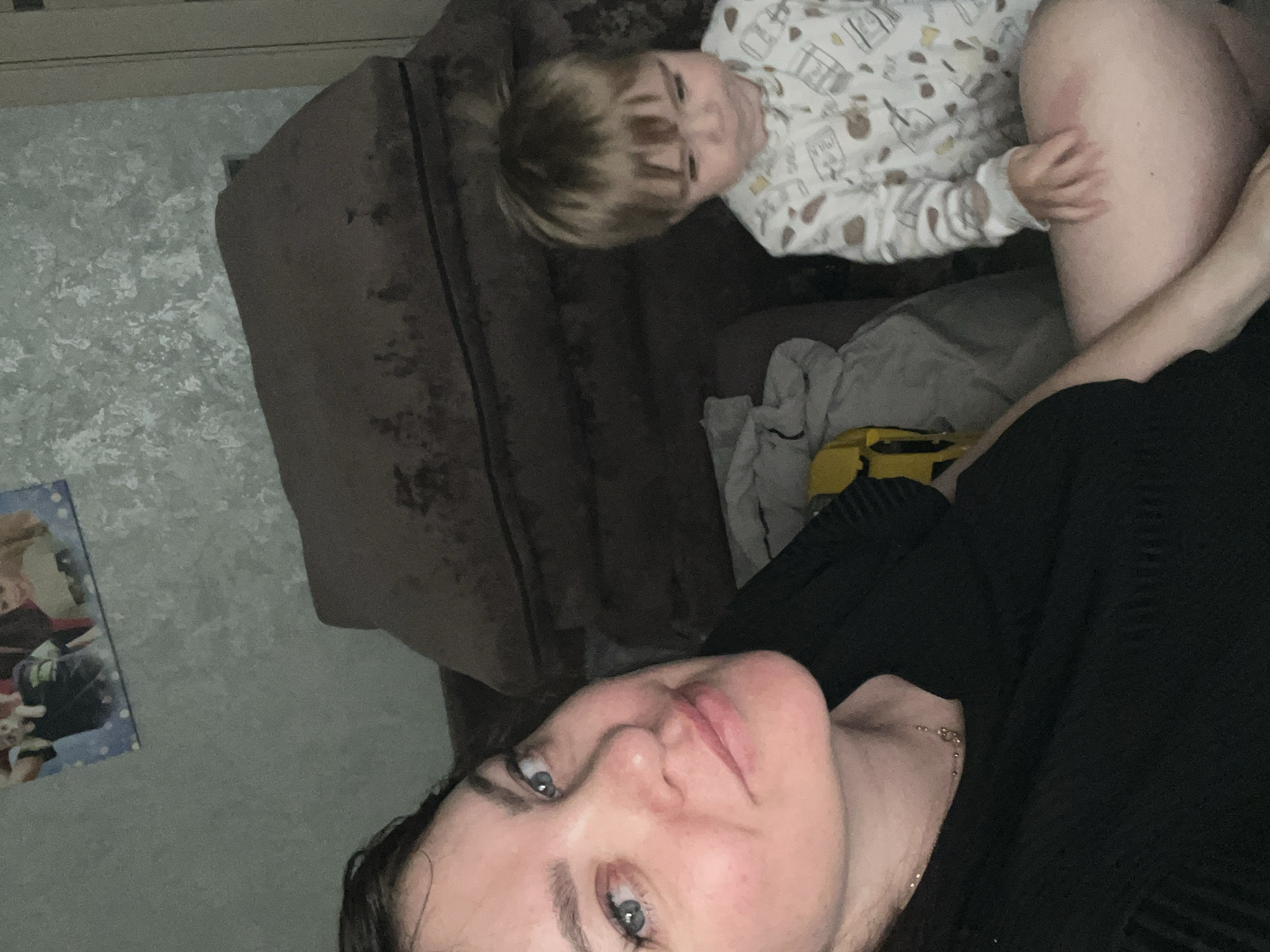 The image depicts an indoor scene in Noviy, Russia, featuring an adult female and a young child. The adult female, positioned in the foreground on the left, has dark hair, blue eyes, and a slight smile, with visible redness on her cheeks and nose. She is wearing a black textured top and a delicate gold-toned necklace, and appears to be taking a close-up photograph from a slightly upward angle.
To her right and slightly behind her, a young child, appearing to be a toddler with light brown hair and bangs, is visible. The child is wearing a white long-sleeved sleepsuit or romper with a pattern of small cartoonish objects (e.g., jars, cups). The child's gaze is directed downwards, away from the camera, and its left hand is resting on the adult female's bare right thigh, where a faint reddish mark is visible.
The background consists of a dark brown upholstered sofa with two visible cushions. On the wall in the upper left, a framed picture or poster displays multiple images of children, including one with a white dog, set against a blue background with white polka dots. The wall in the upper right features a light-colored, textured wallpaper. A folded light-colored blanket or fabric and a partial yellow object are visible on the sofa to the left of the child. A light-colored door frame is partially visible on the far right edge of the image. The scene is illuminated by dim, ambient indoor lighting.