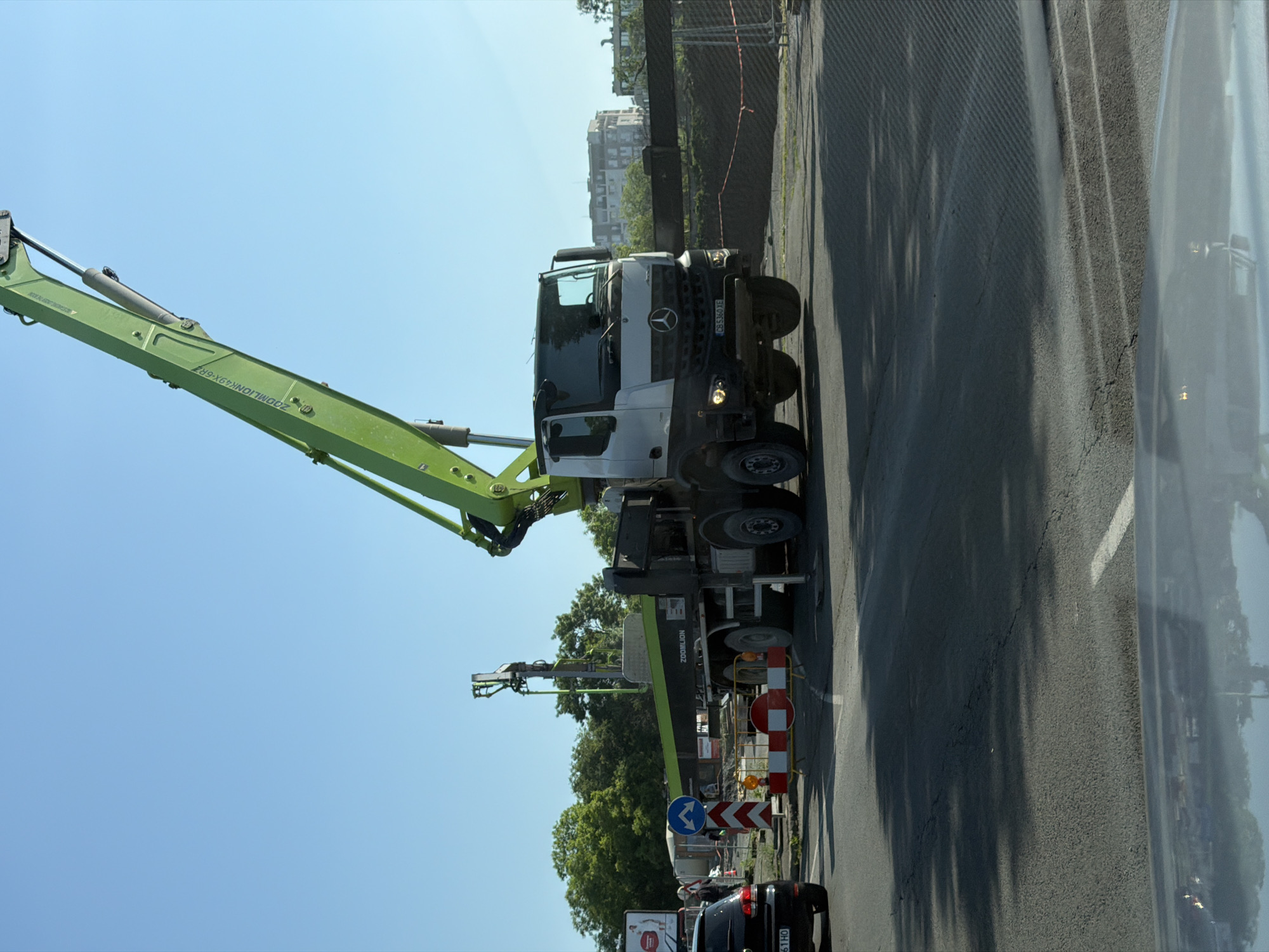 The image, taken in Burgas, Bulgaria, captures a large concrete pump truck positioned on an asphalt road, likely at a construction or roadwork site on a bright, sunny day. The perspective is tilted, suggesting it was captured from a moving vehicle.
The prominent subject is a green and white concrete pump truck. The truck cab is white with a black grille and bumper, featuring the Mercedes-Benz logo on the front. Its license plate, "CB 5360 TE", identifies it as registered in Burgas. Attached to the truck is a long, multi-jointed green pump boom, characteristic of a concrete pump. Text visible on the side of the boom reads "ZOOMLION 49X-6RZ", indicating the manufacturer and model of the pumping mechanism. The boom extends high into a clear, light blue sky.
The truck is situated on a paved road, which shows some cracks and texture. To the right of the truck, there are construction barriers with red and white diagonal stripes, along with a blue circular directional sign showing two arrows curving right and left, indicating a split or detour. Another black car with the license plate "CB 61HO" is partially visible behind these signs, also registered in Burgas. In the background, beyond a line of trees, several residential or commercial buildings can be seen under the clear sky. Shadows of trees or structures are cast across the road surface. The overall scene suggests ongoing infrastructure work.