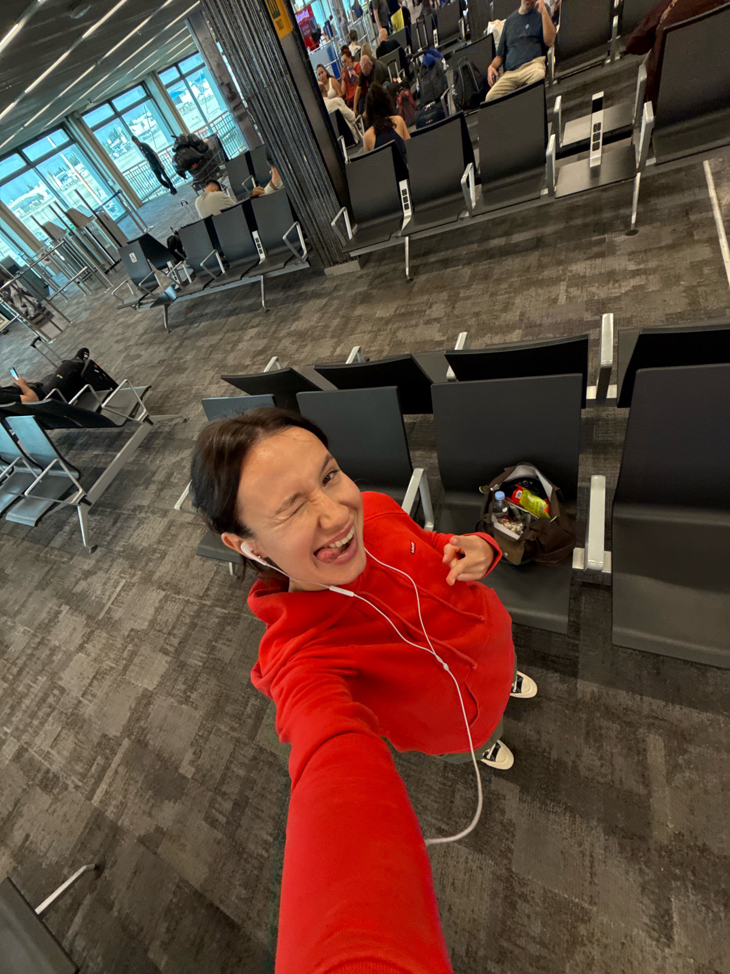 This is a high-angle, full-shot, eye-level, candid photograph of a woman with dark hair and light skin, wearing a bright red hooded sweatshirt and light-colored sneakers. She is in a seated waiting area, likely an airport gate, with rows of black plastic chairs. The woman is facing the camera, winking and sticking her tongue out, with white earbuds in her ears. Her left hand is extended, pointing towards the camera. A brown canvas bag containing snacks and a water bottle sits on a chair next to her. In the background, other people are seated in the waiting area, some with luggage, and large windows offer a view of an outdoor area with vehicles. The floor is carpeted with a textured, gray and brown pattern. The environment appears to be modern and spacious. This scene is identified as being in Yagel, Israel, though the specific location within Yagel is not detailed in the image.