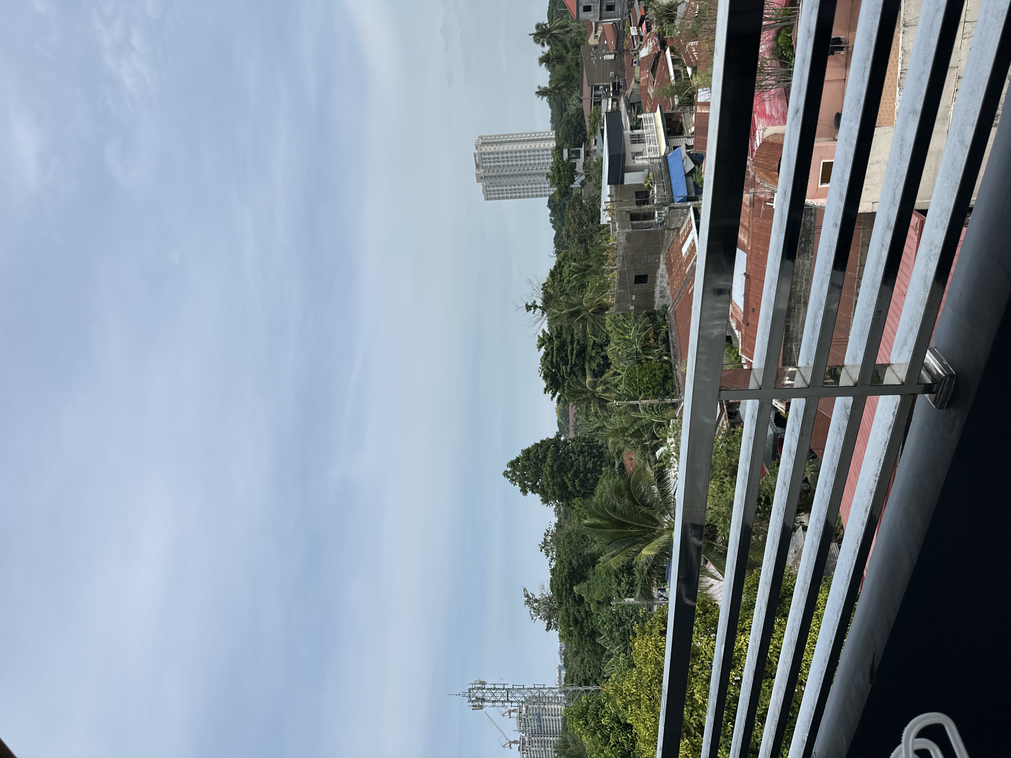 The image shows an elevated view from a balcony in Davao City, Philippines, overlooking a mix of urban and natural environments. The foreground features a grey metal railing. Beyond the railing, the scene includes a variety of rooftops, trees, and buildings. A tall, light-colored building is visible in the distance. The sky is overcast. A cell tower is located in the lower left corner. The image is rotated 90 degrees counter-clockwise. No people are visible.