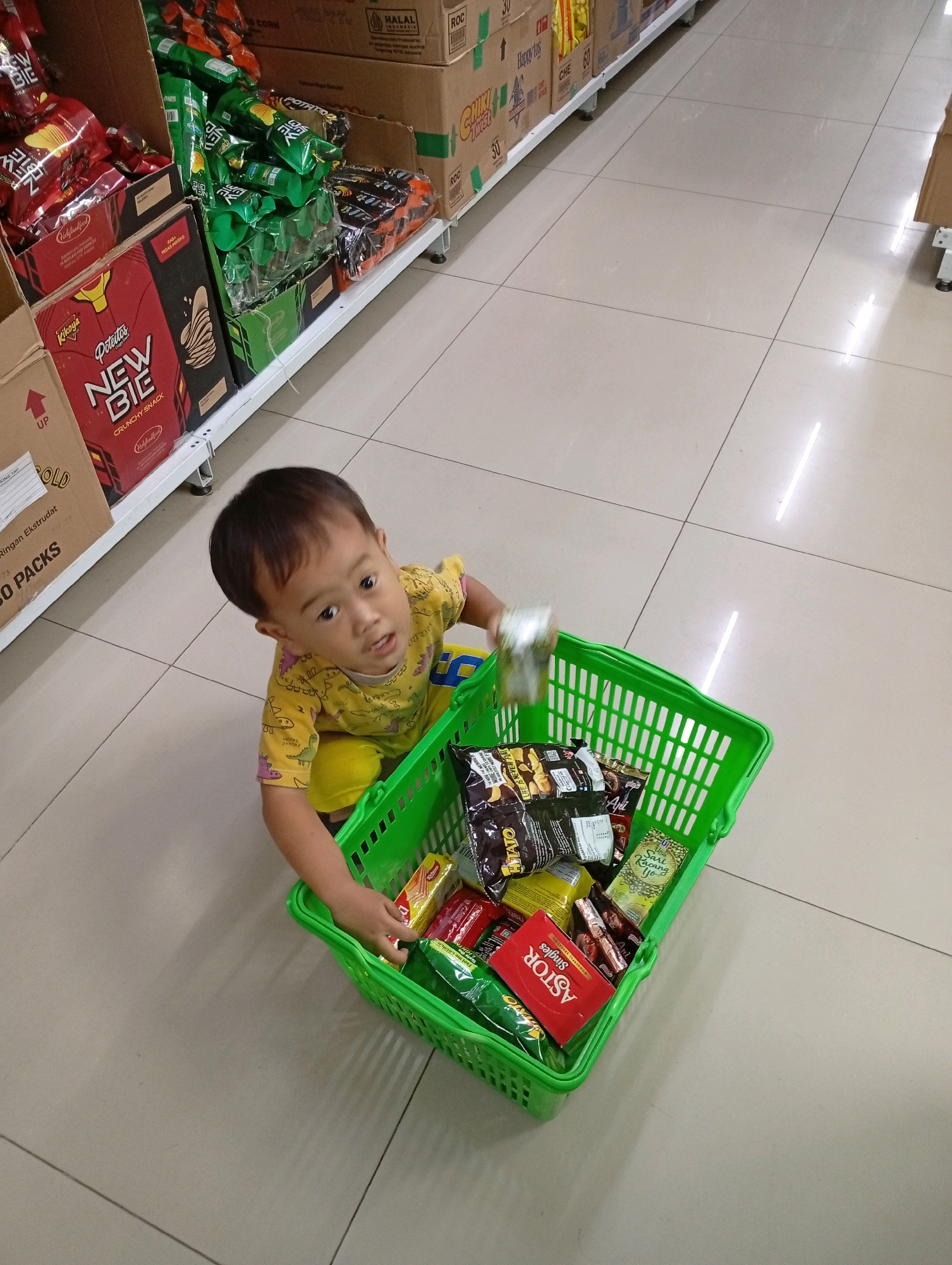 A young Indonesian child, with dark hair and wearing a yellow t-shirt with dinosaur graphics and yellow pants, crouches on a tiled floor in a supermarket aisle in Babakan, Indonesia. The child is looking at the camera and holding a green plastic shopping basket filled with various snack items. The basket contains items such as packaged crackers, biscuits, and other food products. Behind the child, shelves are stacked with more packaged goods, including brightly colored snack bags and boxes. The floor is tiled in a neutral beige color, and the lighting suggests an indoor retail environment. The overall scene captures a child interacting with a shopping basket in a supermarket.