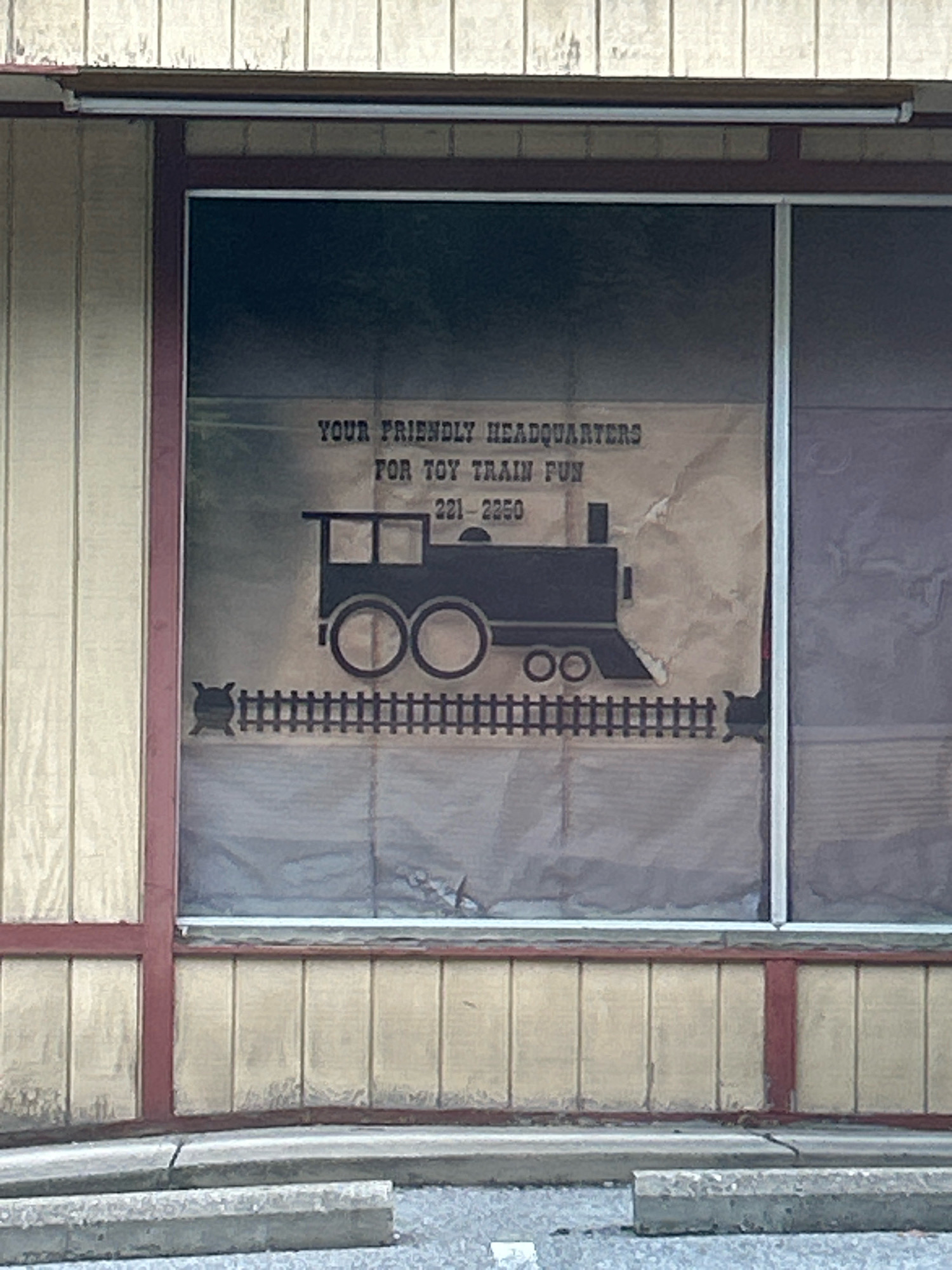 A weathered sign is taped to the inside of a large storefront window, obscuring most of the view of the interior. The sign is beige and features a black silhouette of a toy train with a smokestack and wheels. Above the train, in black, stylized lettering, it reads "YOUR FRIENDLY HEADQUARTERS FOR TOY TRAIN FUN" followed by a phone number, "221 - 2250". The sign also includes a black graphic of a railroad track with a small, dark, stylized locomotive figure at the left end. The storefront itself has vertical siding painted light beige, with dark red trim around the window and along a horizontal band below the window. A metal awning extends above the window. The ground in front of the storefront is paved with asphalt, and there are concrete curbs and a concrete parking block visible. The overall condition of the building and sign suggests a long-standing business. The image does not contain any people or explicit indicators of a specific location within Pittsburgh, United States of America, other than it being a commercial establishment.