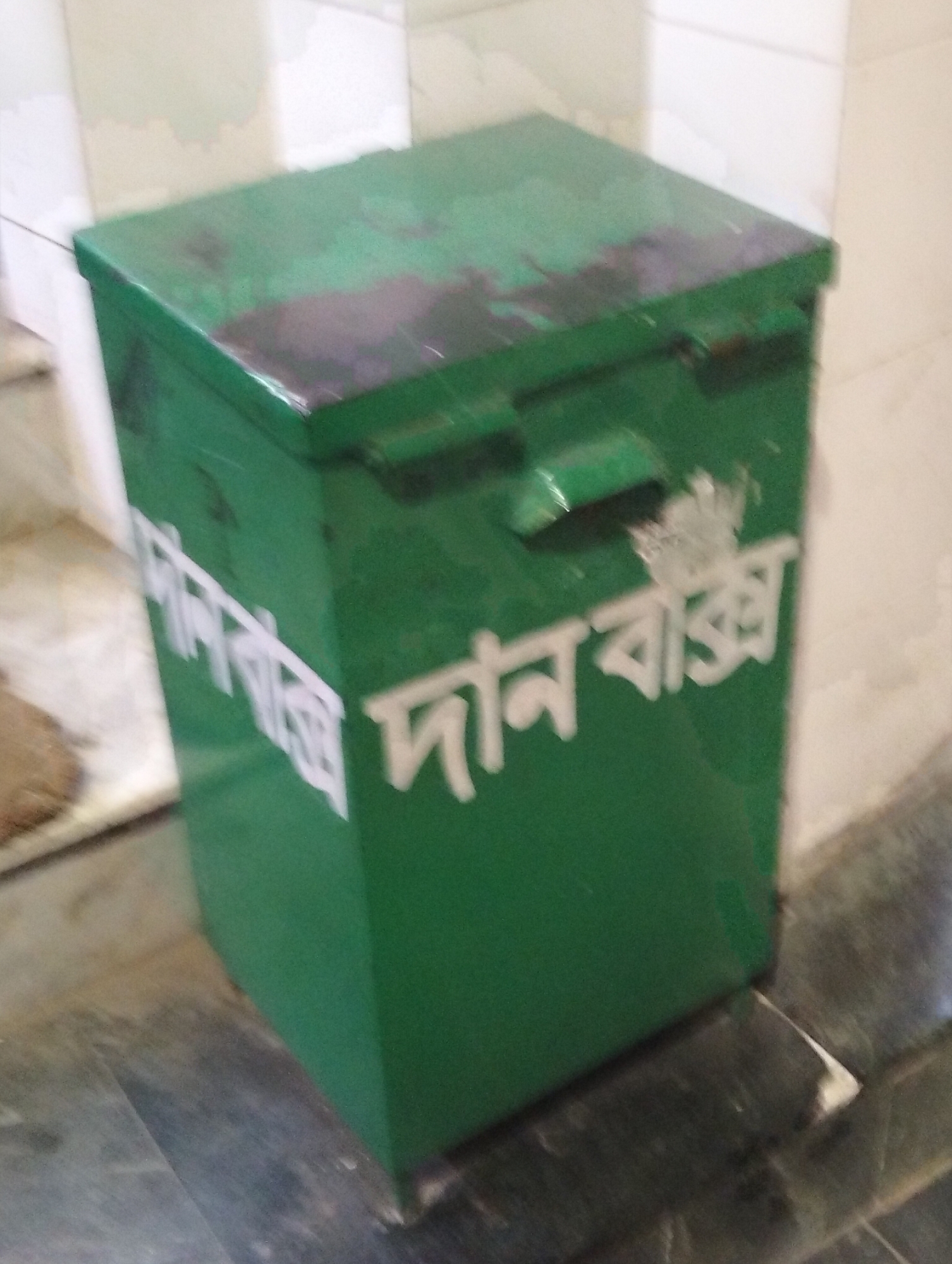 A medium shot depicts a green metal donation box with white Bengali script that translates to "Donation Box." The box is rectangular and shows signs of wear with some darker spots. It has a handle and is positioned on a dark, possibly marble-like, floor. The background includes a tiled wall that appears to be in a building interior. The location is presumed to be Dhaka, Bangladesh, based on the script on the box.