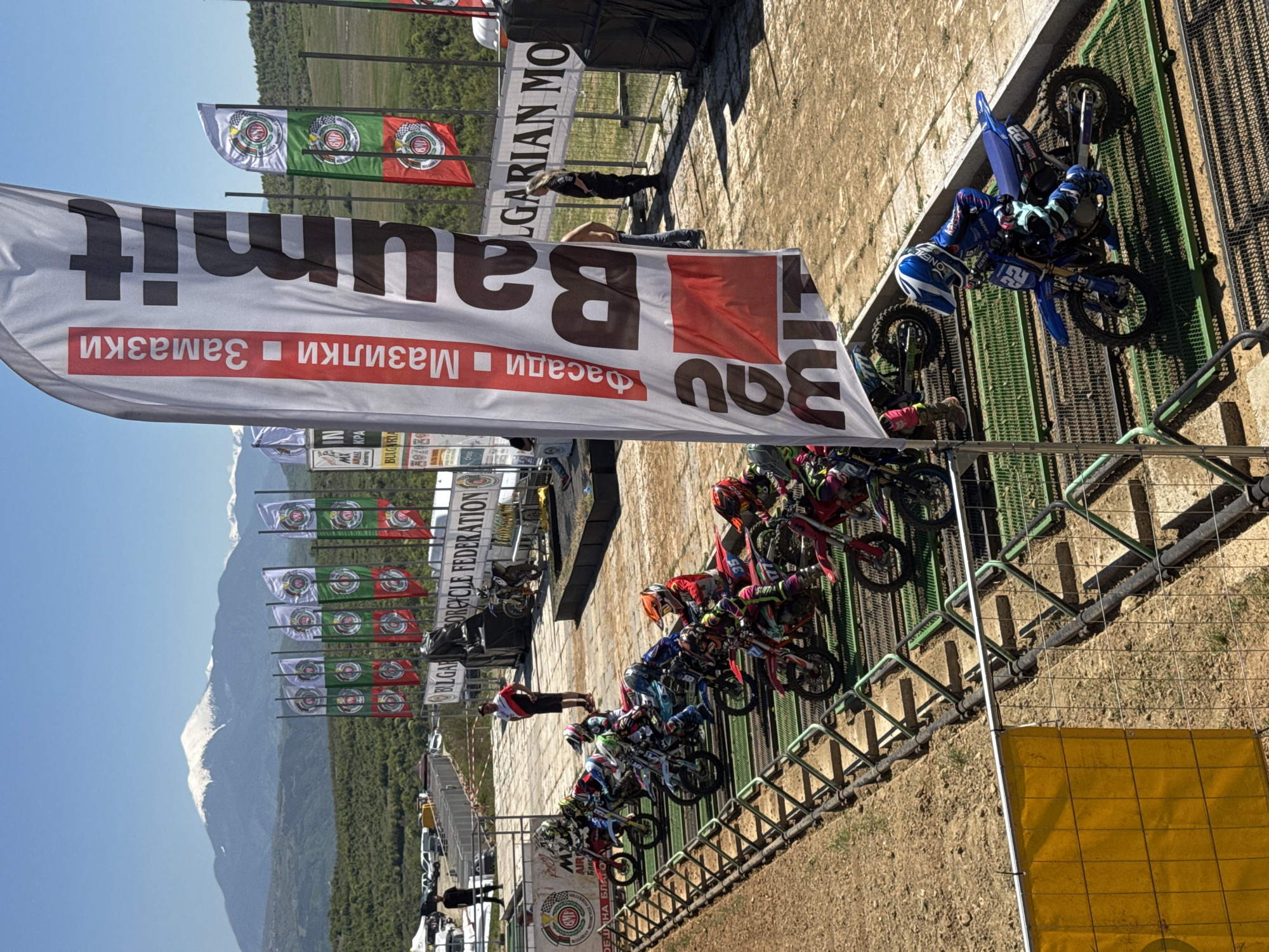 The image shows a motocross event, possibly at the beginning of a race. There are several riders on their motorcycles lined up behind a starting gate. Large banners with the "Baumit" logo are visible, as well as flags representing the "Bulgarian Motorcycle Federation". A mountain is seen in the background. The event is taking place in Selishte, Bulgaria. Several people are standing near the starting gate. The motocross riders are wearing helmets and protective gear.