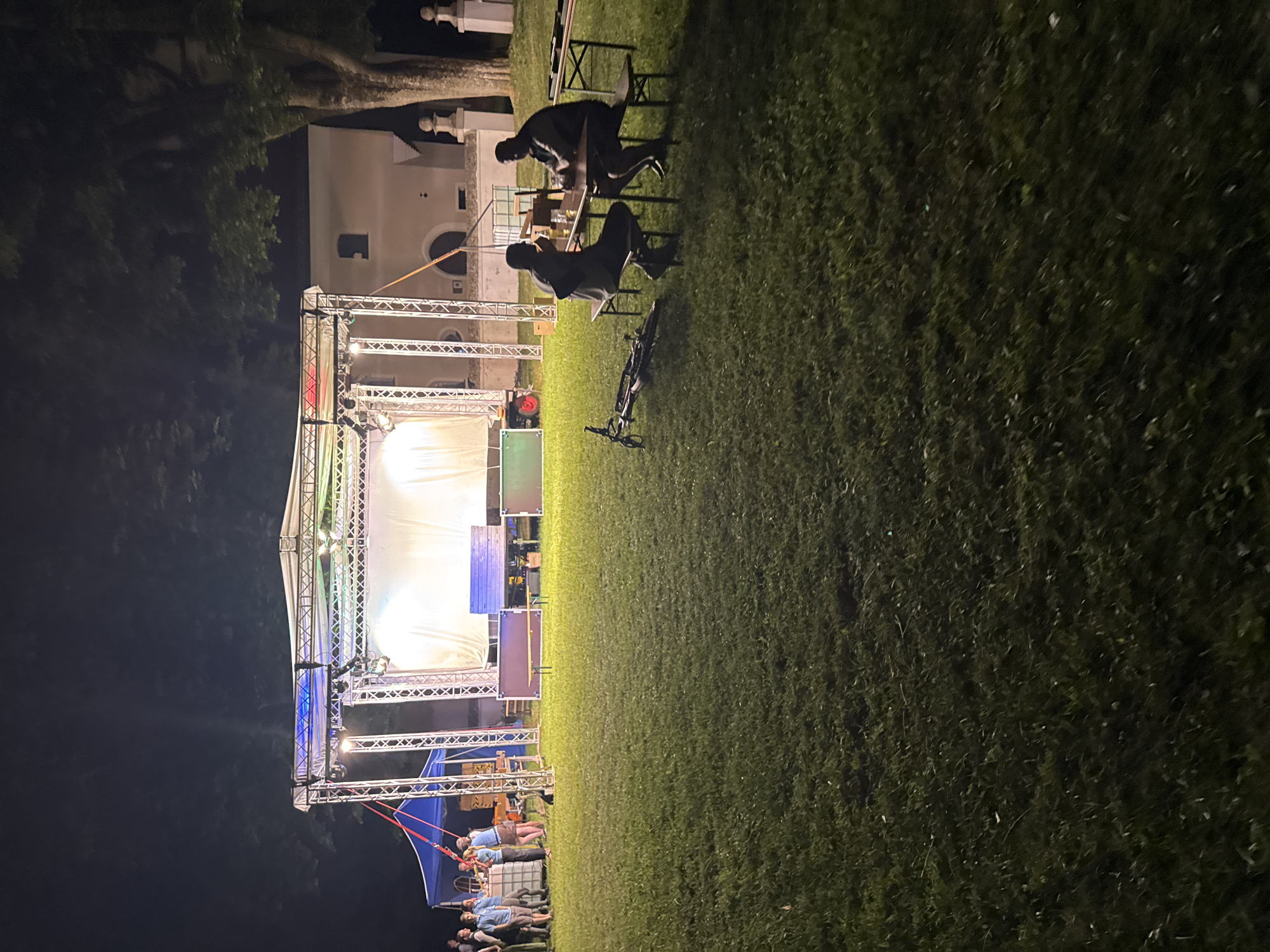 An outdoor event is taking place at night on a grassy field in Höhenkirchen-Siegertsbrunn, Germany, with a stage set up in the background. The stage has lights illuminating a white screen and is supported by a metal truss structure. A building with arched windows and a tower is visible behind the stage. On the left side of the image, a blue tent is set up, and a group of people are gathered near it. In the foreground, two men are seated at a picnic table, with drinks on the table. A bicycle is lying on its side on the grass in front of them. The field is covered in green grass, and trees are visible in the background.