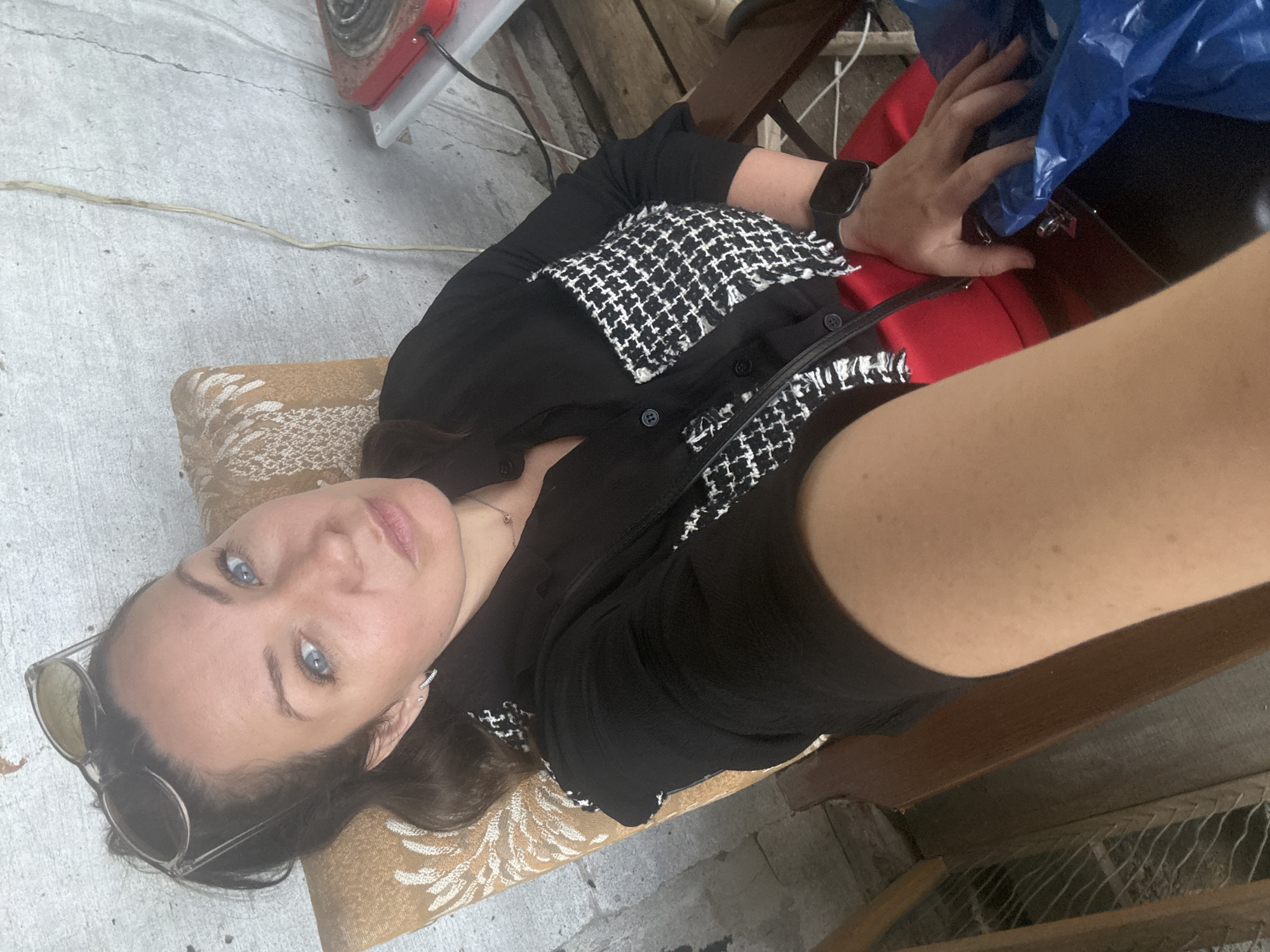 A woman with blue eyes and dark hair pulled back is seated in a chair and taking a selfie. She is wearing sunglasses on her head, a black shirt with white trim, and a red skirt. A watch is visible on her left wrist. She is sitting in what appears to be an outdoor or semi-outdoor setting, with a concrete wall behind her and wooden flooring beneath. To the right of the frame, a red electric hot plate is visible on a white shelf. The overall environment suggests a simple, utilitarian space, possibly an outbuilding or a workshop. The image's lighting and subject matter do not provide specific clues to its location within Chaikovsky, Russia.