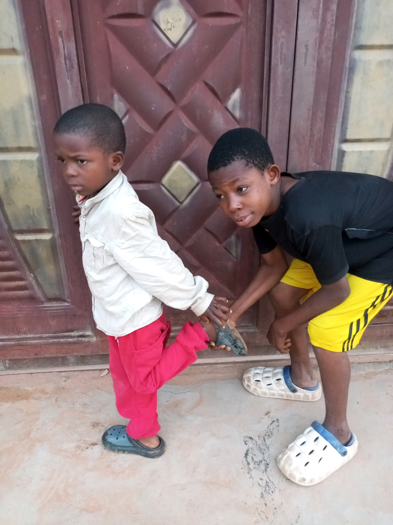 Two young boys are standing in front of a dark, patterned door. The boy on the left is wearing a light-colored collared jacket and red pants. He is lifting one leg, and the boy on the right is holding his hand. The boy on the right is wearing a black t-shirt and yellow shorts with black stripes. He is crouching and looking at the other boy. Both boys are wearing clogs on their feet. The ground is made of concrete.