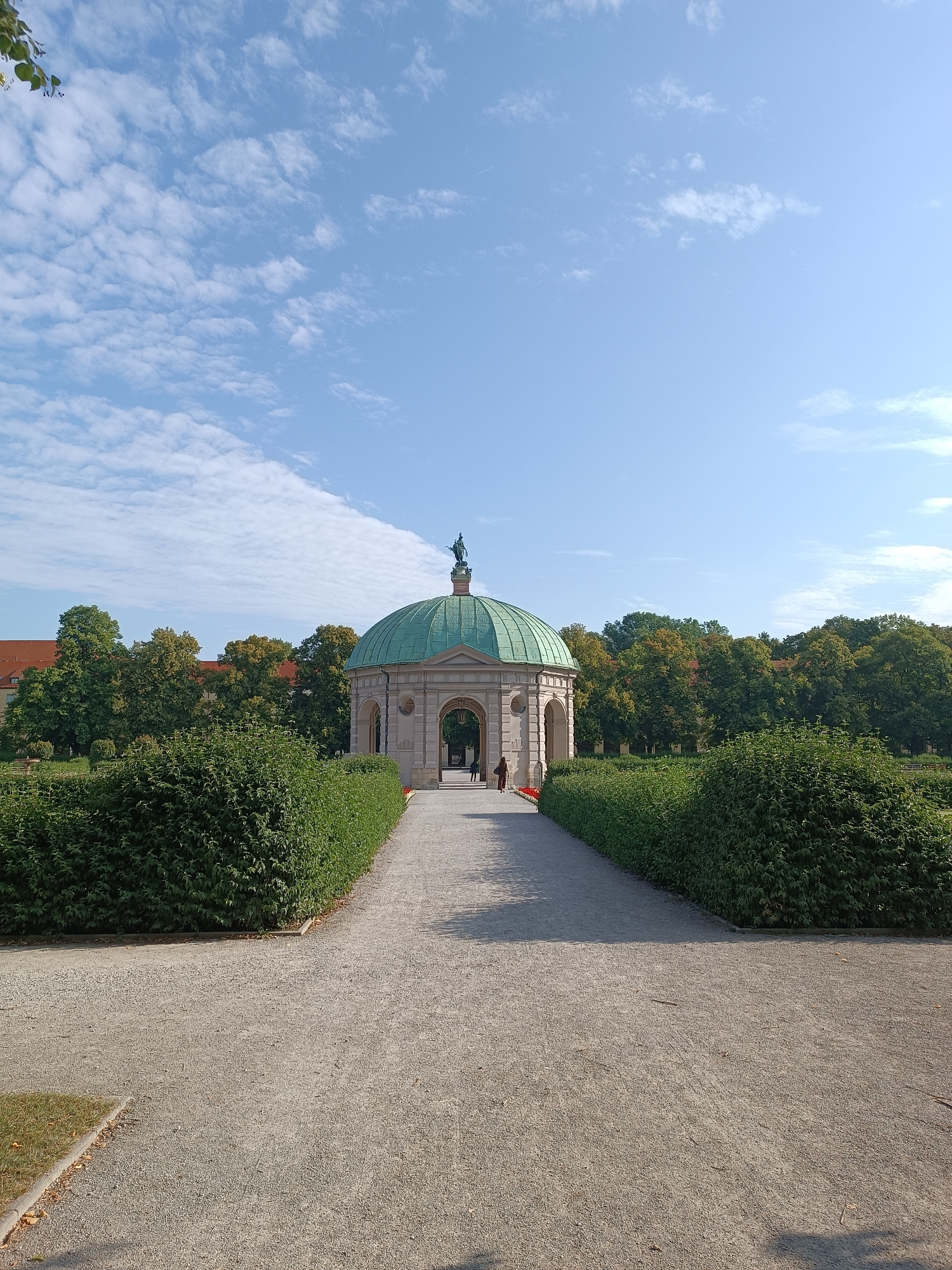 This is a photograph of the Monopteros in the English Garden in Munich, Germany. The Monopteros is a circular temple with a green copper dome, supported by columns. A statue of Apollo stands atop the dome. The temple is situated on a small hill, surrounded by manicured hedges and gravel pathways. In the background, a line of trees with green and orange foliage is visible, and beyond that, the reddish-brown roof of a building can be seen. Two individuals are walking towards the temple from the left side of the frame. The sky is blue with scattered white clouds, and sunlight casts shadows on the ground.