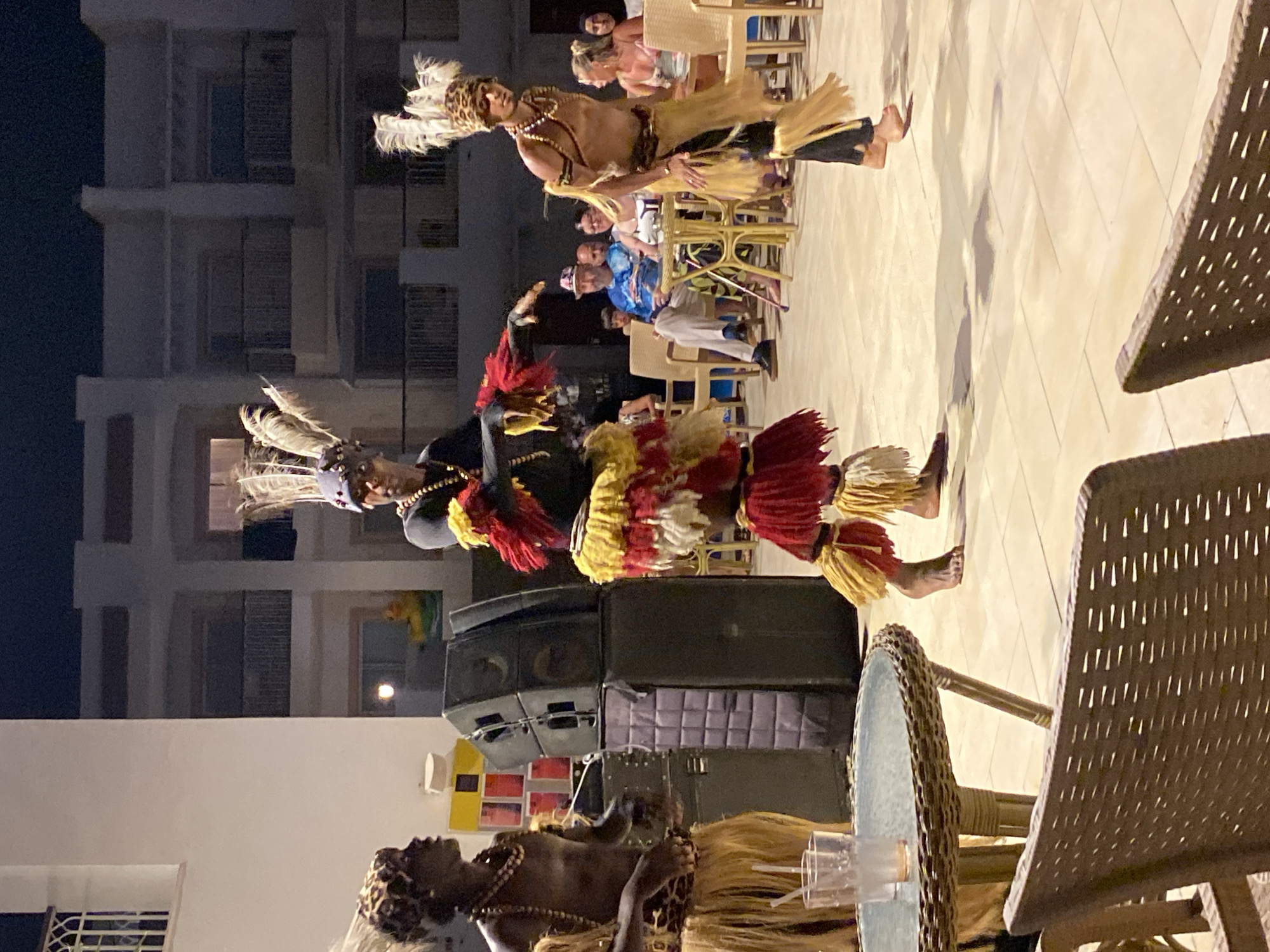 In Sharm El Sheikh, Egypt, at night, three performers in traditional attire with feathered headdresses are dancing on a tiled outdoor patio. Two are in the foreground, one on the left facing left and the other in the center facing right with a headdress adorned with feathers and leopard print. A third performer is on the right, facing left, also wearing a feathered headdress and a skirt made of straw-like material. The performers have elaborate headdresses with feathers and wear beaded necklaces and tribal-style skirts. In the background, a multi-story building with balconies and illuminated windows suggests a resort or hotel. Several people are seated at tables in the middle ground, observing the performance. Speakers are stacked behind the performer on the left. In the foreground, a round wicker table with a glass top holds two drinks. The lighting is artificial, creating shadows on the patio.