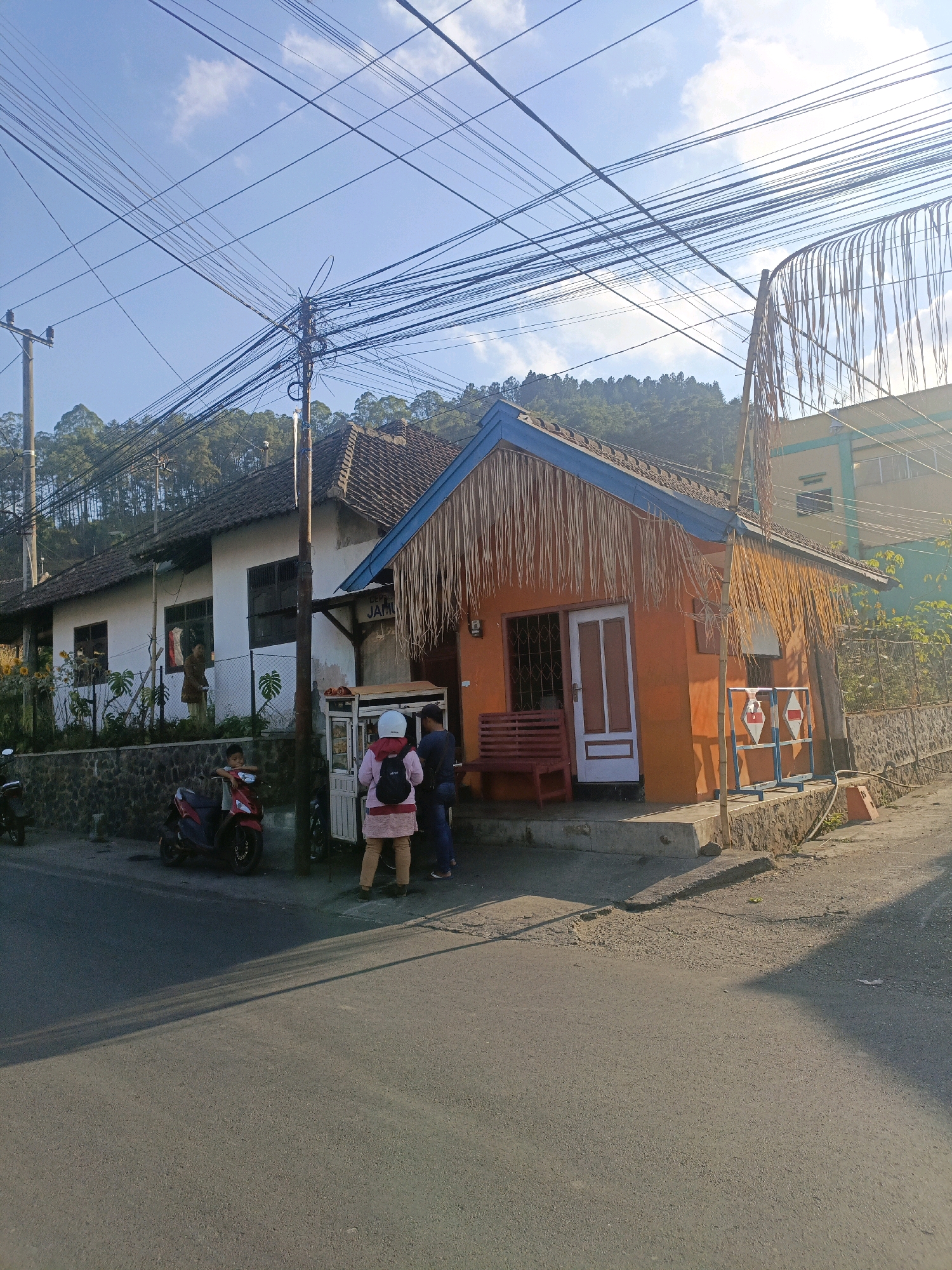 In Batu, Indonesia, on a sunny day, a street scene unfolds with a small, vibrant orange building featuring a thatched roof and blue trim. To its left, a larger, white building with a dark tiled roof is partially visible. In front of the orange building, a red bench sits outside. Two people, one wearing a pink top and white helmet, and another in a dark shirt and pants, stand near a mobile food cart. A red motorcycle is parked to the left of the food cart, with a young boy sitting on it. The street is paved, and a steep, tree-covered hillside rises behind the buildings, crisscrossed by a dense network of overhead electrical wires. The overall atmosphere suggests a rural or semi-urban setting with active local commerce.