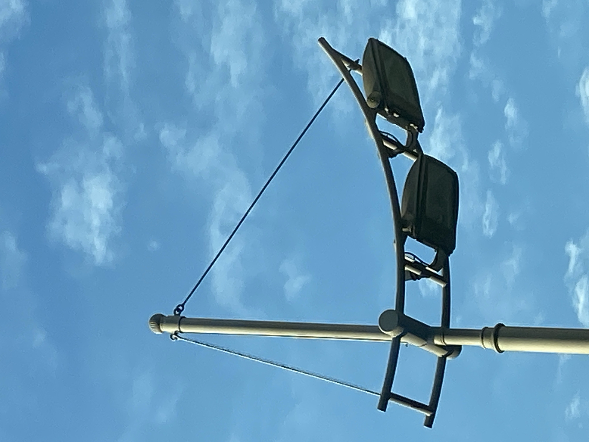 A street lamp with two lamps is mounted on a pole against a blue sky with white clouds. The street lamp is made of light-colored metal and has a curved arm from which the two lamps are suspended. The lamps are dark and angled downwards. The pole is also made of light-colored metal and has cables attached to it. The scene appears to be in Budva, Montenegro.