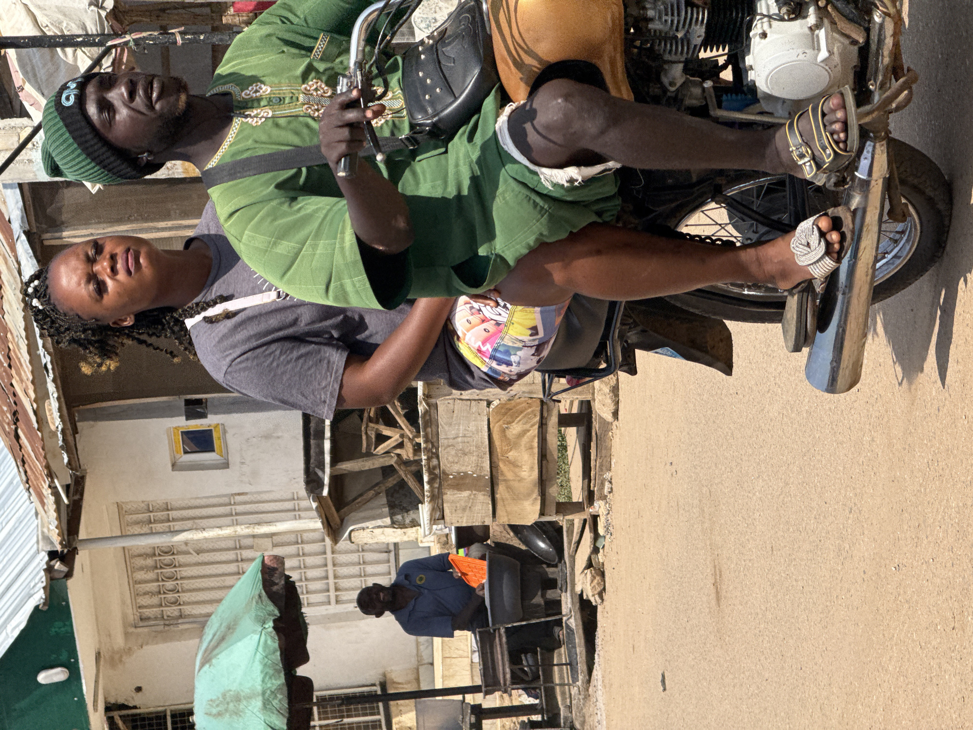 Two individuals, a man and a woman, are seated on a motorcycle in an outdoor, urban environment. The man is in the front, operating the motorcycle, and is wearing a green dashiki-style shirt, green shorts, and a green beanie with a black band. He has a black crossbody bag with metal studs slung over his right shoulder. The woman is seated behind him, wearing a grey t-shirt and brightly colored shorts.
The motorcycle is yellow and brown. In the background, a man in a blue shirt is visible, sitting near a stall and holding an orange object. The environment appears to be a market or street with simple structures, some with corrugated metal roofs. A green umbrella is also visible in the background. The ground is a dusty, light brown. The overall scene suggests a typical day in a bustling Nigerian city, potentially in Abuja, given the context.