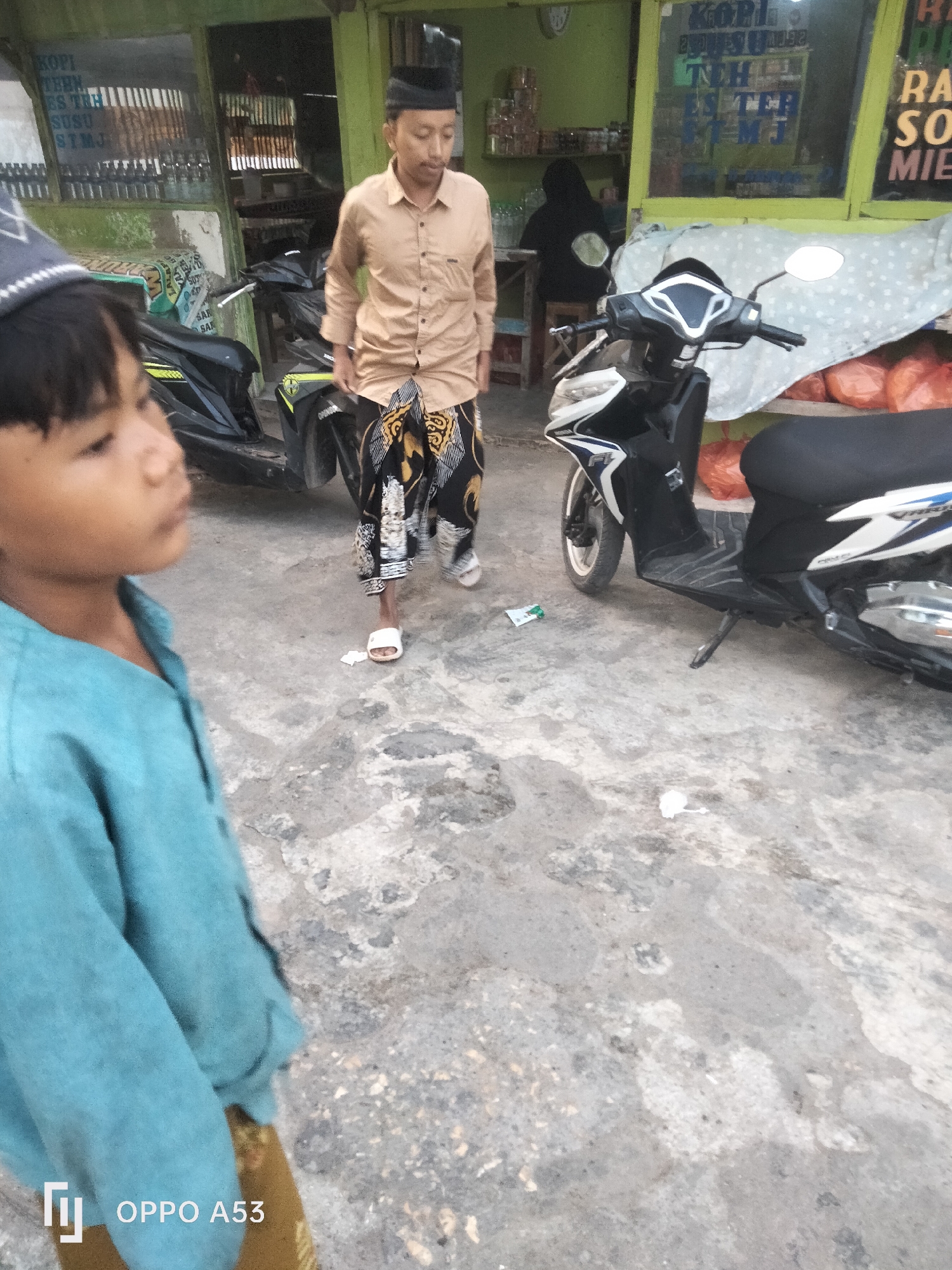 The image depicts a street scene in Badung, Indonesia, with a focus on a young child in the foreground and a man in traditional clothing in the midground. The child, wearing a teal shirt, is looking to the right, out of frame. The man, dressed in a tan shirt and a patterned sarong, is standing near two motorcycles. One motorcycle is black and white, and the other is black. Behind the man, a shop with a green facade is visible, displaying various goods. The ground is paved with concrete. The lighting suggests it is daytime, possibly late afternoon, given the shadows. The overall environment appears to be a commercial or residential area typical of a bustling Indonesian city.