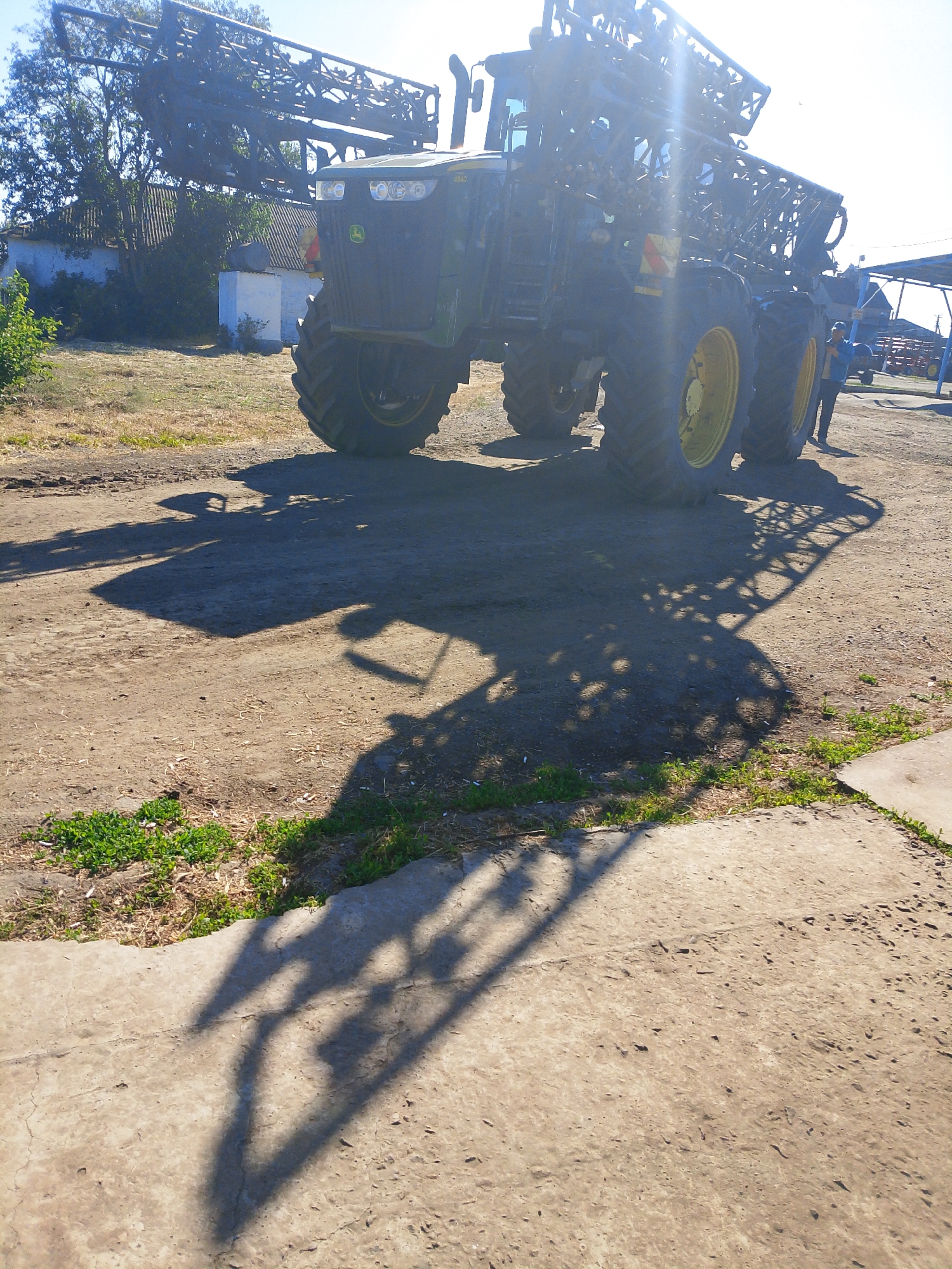 In Dobra Krynytsia, Ukraine, a large, green agricultural sprayer, identified as a John Deere 4240, is parked outdoors on a dirt and concrete surface. The sun is shining brightly, casting long shadows from the sprayer and the surrounding environment. The sprayer has a massive boom assembly, characteristic of large-scale agricultural equipment. In the background, a white building with a striped roof is visible, along with trees and vegetation. A person, wearing a blue shirt and a cap, stands near the rear of the sprayer. The ground is uneven, with patches of dry grass and scattered debris. The overall scene depicts a typical rural setting, likely on a farm.