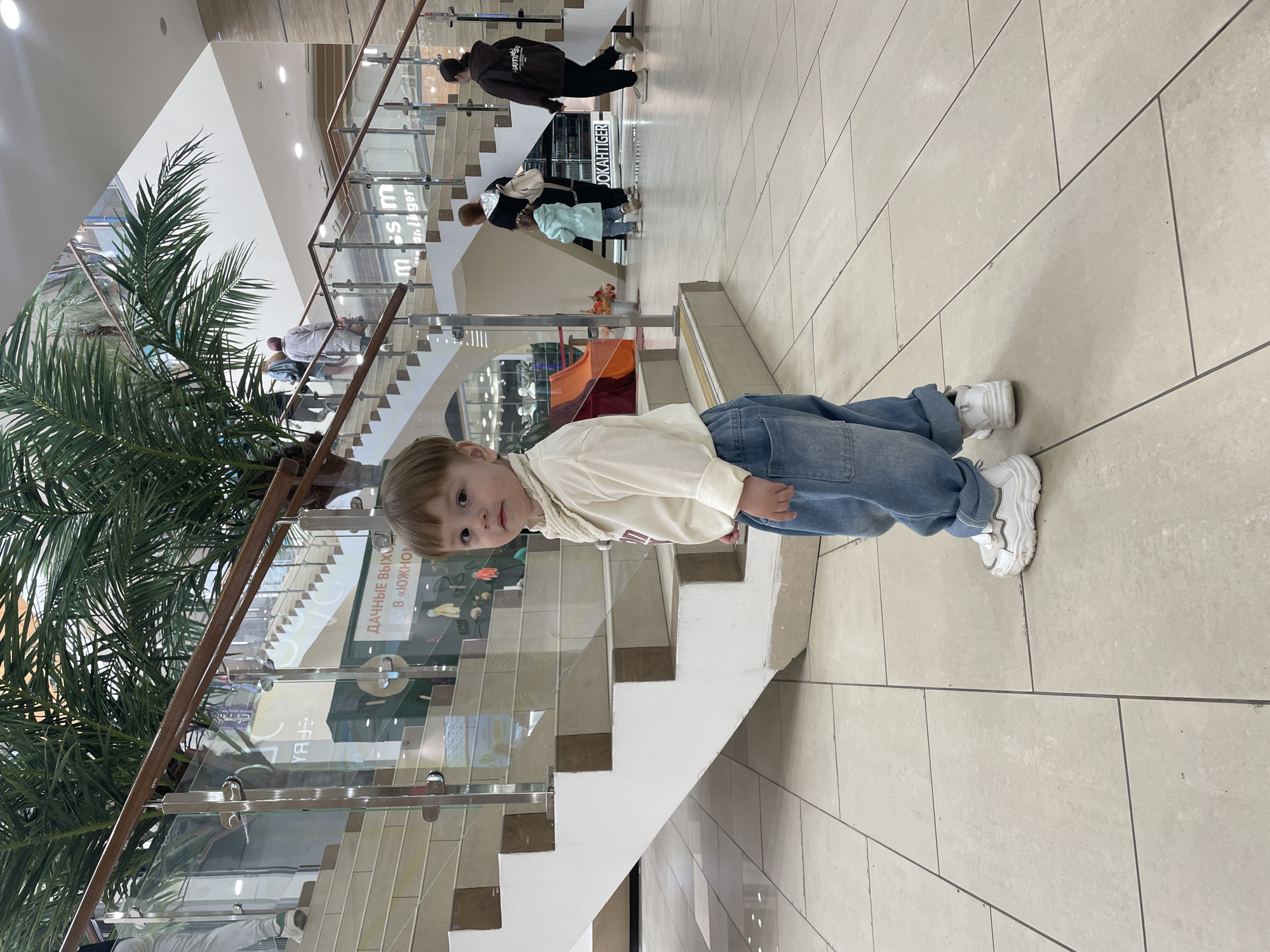 The image depicts a young child, appearing to be around 3-5 years old, lying prone on a tiled floor, seemingly in a shopping mall. The child is wearing a light-colored top, denim jeans, and white sneakers. The mall environment is characterized by multiple levels with glass and metal railings. A large potted palm tree is visible on one side. The stairs near the child have beige and white tones. In the background, blurred figures of people can be seen walking on different levels of the mall. The text on the glass railing is in Russian and likely pertains to store names or advertising. Based on the context and text on the storefront, the image is likely taken at the Mega shopping mall in Kazan, Russia.