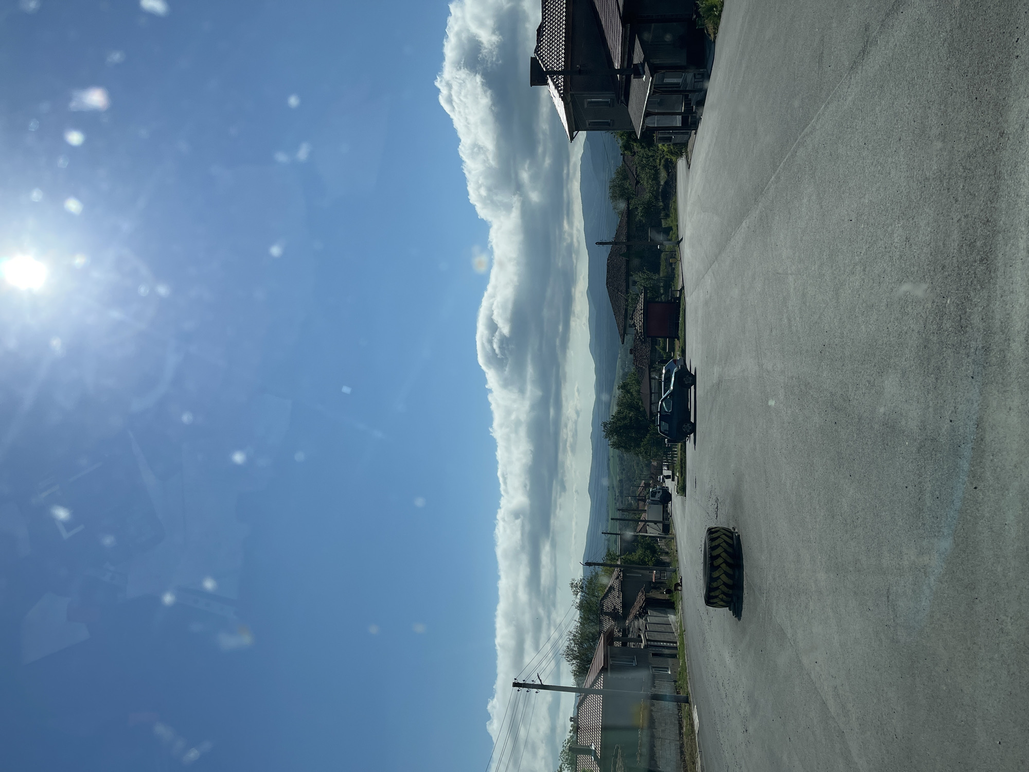 A sunny day in Rish, Bulgaria. The image is taken from inside a vehicle, looking out onto a street. A large tire is in the middle of the road. Houses line one side of the street, with power lines visible. Mountains are visible in the distance behind the houses. Cumulus clouds are on the horizon. The sky is blue with a bright sun on the left.
