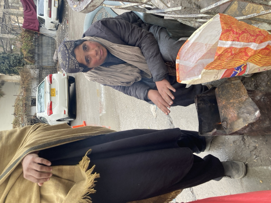 The image depicts an outdoor urban scene in Quetta City Tehsil, Pakistan, characterized by damp ground and an overcast environment.
In the foreground, two individuals are present. On the left, a person stands, partially visible from the mid-thigh up. They are wearing a light brown fringed shawl over dark-colored baggy trousers and grey shoes. Their right hand is visible, positioned at waist level.
To the right, a second individual, appearing youthful, is seated on a light blue plastic chair. This person wears a dark, intricately patterned cap, a dark padded jacket, a light grey scarf wrapped around the neck, and dark trousers. Their hands are clasped together and held directly above a makeshift brazier, constructed from a rusted, cut-open metal drum, which contains a small, visible fire. The individual is looking directly towards the viewer.
Adjacent to the brazier, on the ground, a large, upright shopping bag with an orange, red, and white patterned design, bearing the word "PAKISTAN" and other text, is visible.
In the background, on a paved surface, two white sedans are parked. The closest is a Toyota Corolla with the Balochistan license plate "BQT-163." An orange traffic cone is positioned near this vehicle. Behind the vehicles, a building with a dark metal fence and some sparse, dry vegetation is visible. The ground throughout the visible area appears wet and dirty.