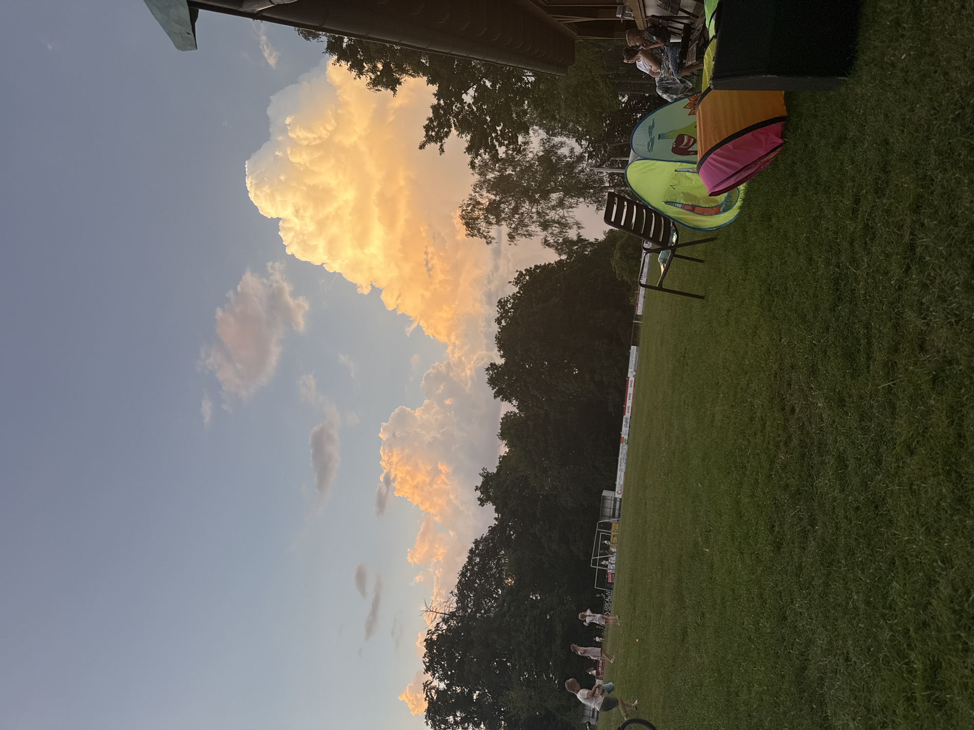 The image depicts a park-like area in Lumda, Germany, likely during late afternoon or early evening. The sky is a pale blue with dramatic, brightly lit cumulonimbus clouds in shades of orange and yellow, indicative of sunset or a storm. In the foreground, a grassy field is visible, with several children playing in the distance. Some adults are seated near a building structure on the right side of the frame. Scattered on the grass are children's play tents and chairs, suggesting a recreational gathering. The background is dominated by a dense line of trees. The overall atmosphere suggests a relaxed outdoor event.
