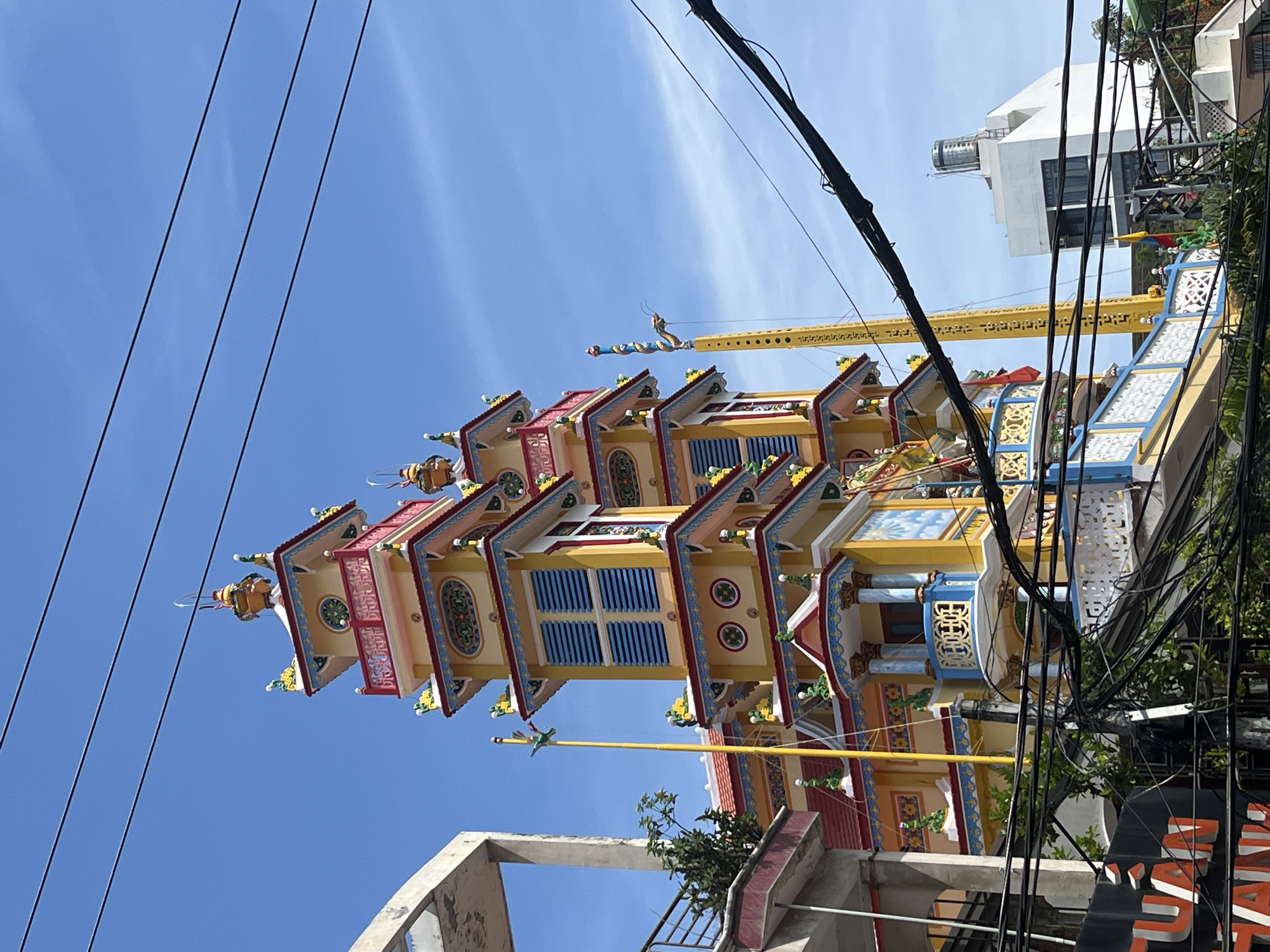 The image captures a vibrant, multi-tiered pagoda structure in Nha Trang Ward, Vietnam, against a clear blue sky. The pagoda is predominantly yellow with red and blue accents, adorned with decorative elements and ornate railings. Several tall, thin poles with flags or streamers are visible in front of and beside the pagoda. In the foreground, a dense network of black electrical wires crisscrosses the frame, obscuring parts of the lower sections of the buildings. To the left, a concrete structure with a balcony and some greenery is partially visible. To the right, a modern white building with a water tank on its roof stands further in the background. The overall impression is a juxtaposition of traditional religious architecture with the utilitarian infrastructure of a modern urban environment.
