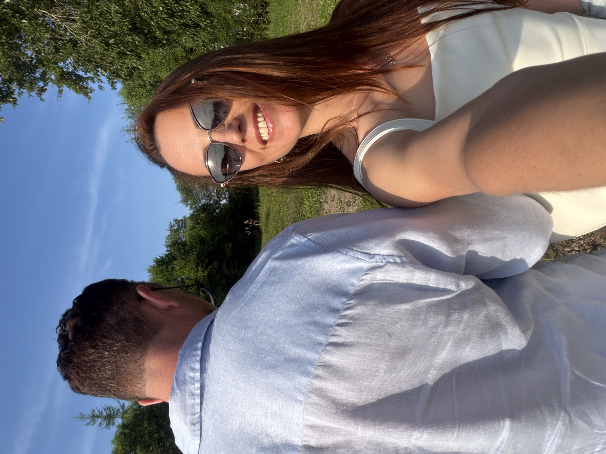 The image shows a selfie taken outdoors, featuring a young woman and a man. The woman is in the foreground, smiling at the camera. She has long, reddish-brown hair, is wearing sunglasses, and a white dress or top. The man is visible behind her, his back turned to the camera, wearing a light blue collared shirt. They appear to be in a natural setting with trees and green grass in the background. The sky is blue with some clouds, suggesting it is daytime with clear weather. The overall mood appears cheerful and relaxed, possibly a casual outing or a special occasion. The image is oriented vertically. There is no visible text in the image.