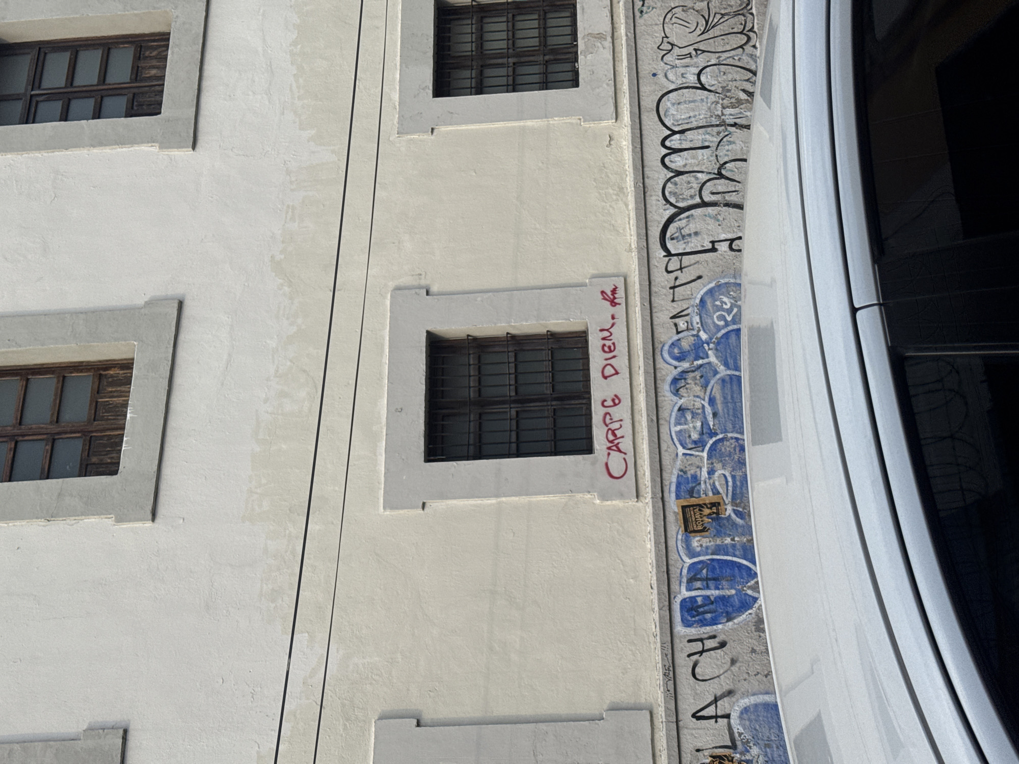 The image depicts a section of a building facade in Puebla, Mexico. The building is characterized by multiple windows, some of which have dark metal bars or grilles. A distinct linear element runs vertically along the building's surface. Graffiti is present on the building's exterior, including a phrase that reads "Carpe Diem." A white vehicle is parked near the building, partially obscuring the view of the graffiti.