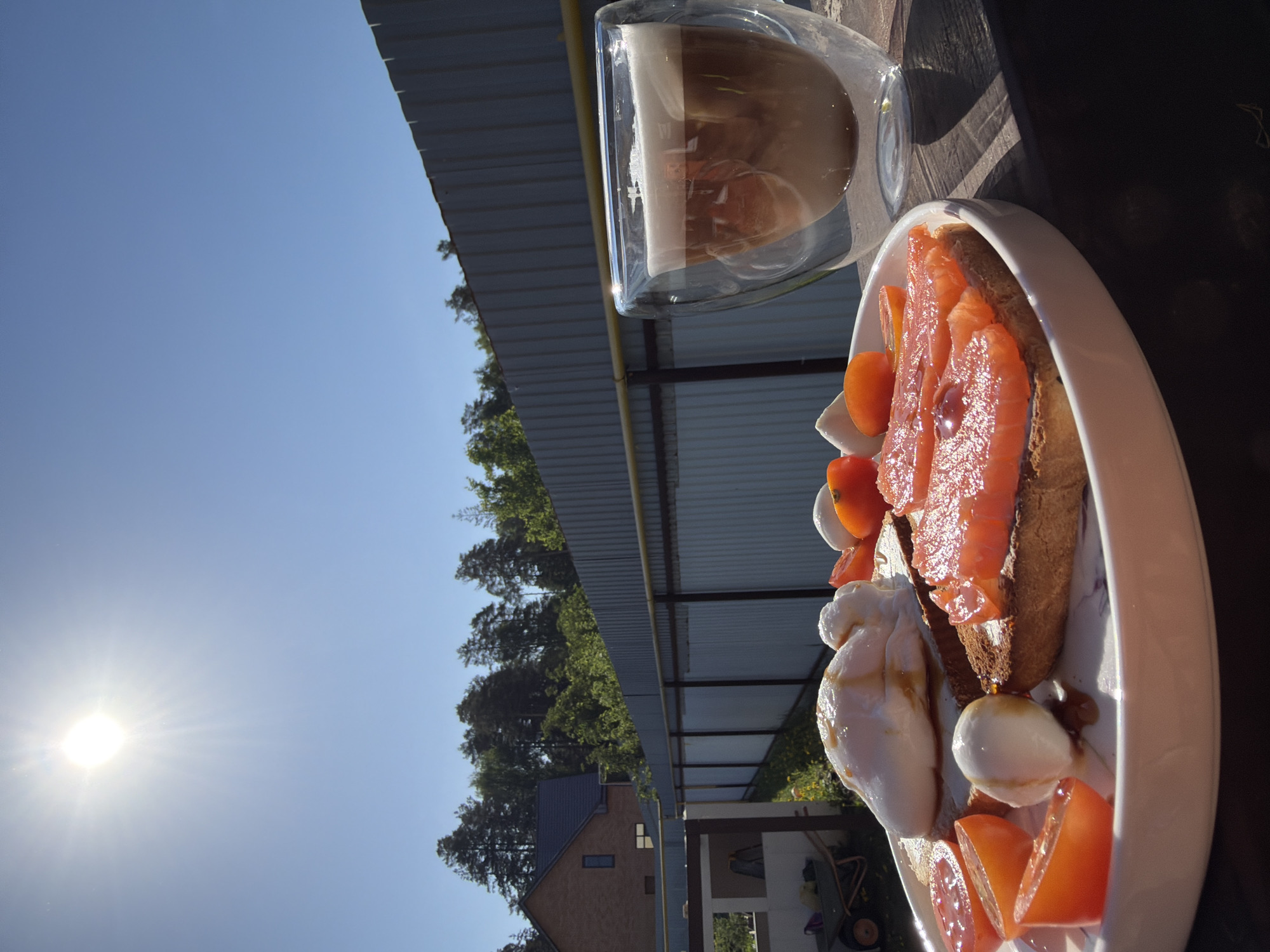 A plate of food and a glass of latte are on a wooden surface under a metallic structure. The plate contains sliced tomatoes, bread, smoked salmon, and mozzarella cheese. Outside the metallic structure, there is a clear blue sky and a building surrounded by trees. The location is implied to be in Chaikovsky, Russia.