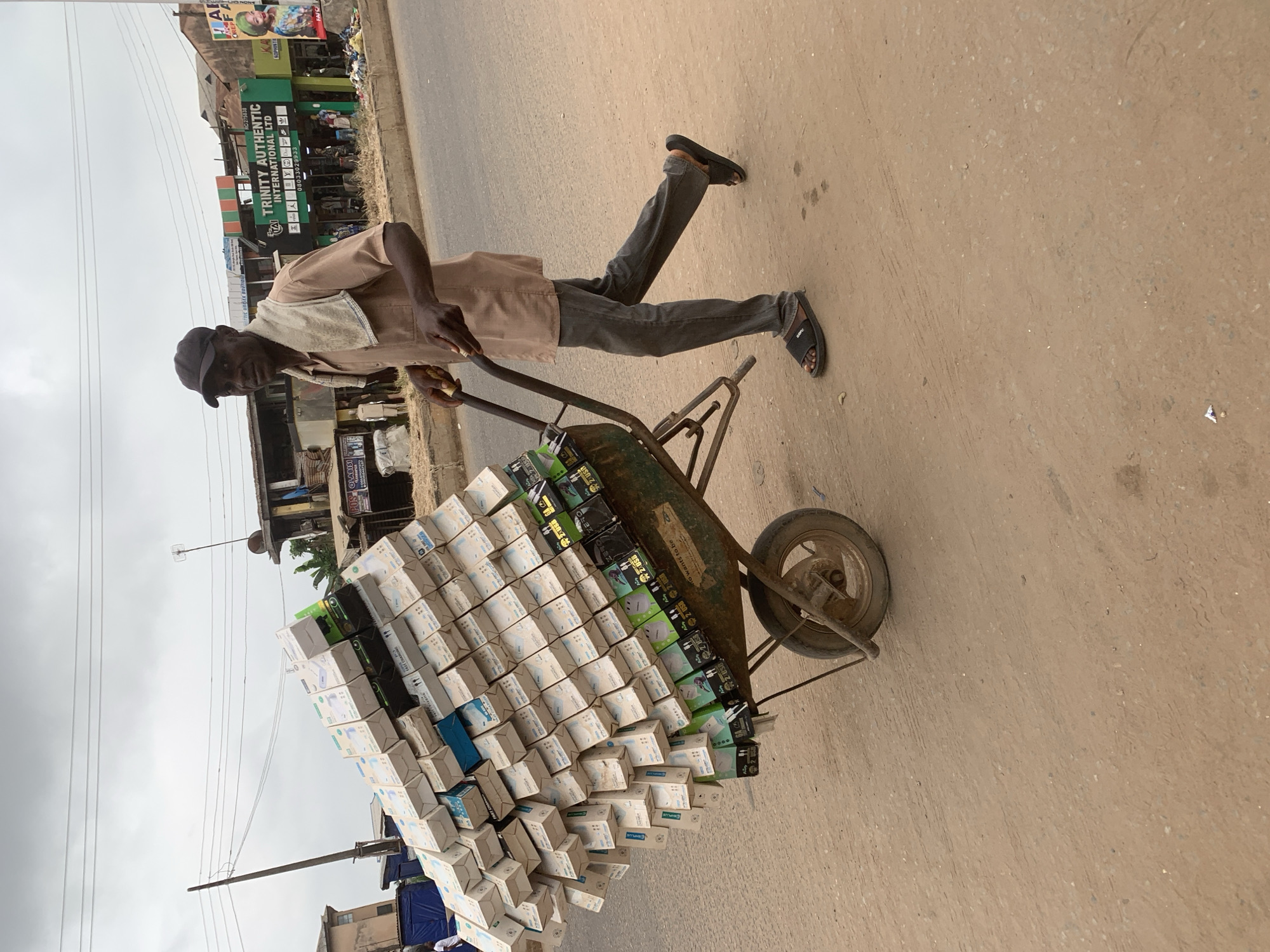 In Shagamu, Nigeria, a man pushes a heavily laden wheelbarrow down a dusty road. The wheelbarrow is piled high with numerous white and blue boxes, likely containing electronic accessories such as phone chargers, as indicated by labels like "FAST CHARGER" and "2 USB" visible on some of the lower boxes. The man, wearing a light brown short-sleeved shirt, dark grey trousers, a black baseball cap, and black sandals, is captured mid-stride, with his right hand gripping one of the wheelbarrow's handles. A light-colored towel is draped over his shoulders. Behind him, a street scene unfolds with several commercial buildings, including "TRINITY AUTHENTIC INTERNATIONAL LTD," a shop with green and orange signage, and other establishments with various posters and signs. Overhead, electrical wires crisscross the cloudy sky, and in the background, a few banana trees are visible, suggesting a tropical climate. The road surface is unpaved and appears to be a dirt or laterite material, with some scattered debris. The overall impression is one of urban commerce in Nigeria, with a focus on the logistics of moving goods via manual labor.