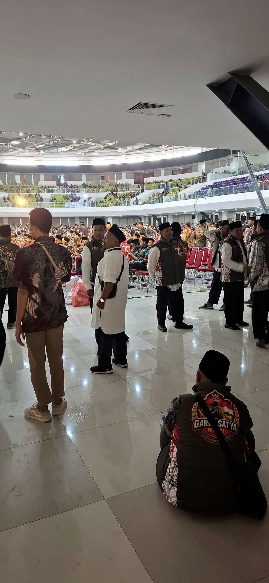 The image depicts an indoor gathering, possibly a convention or a meeting, within what appears to be a large hall or stadium. In the foreground, several individuals are visible, some standing and others seated. One person in the center foreground is wearing a white long-sleeved shirt and black pants, with a black bag slung across their body. To their left, another person wears a maroon patterned shirt and tan pants. Further into the scene, a group of men in black caps and vests, some with insignia on their vests, are engaged in conversation. In the background, tiered seating filled with people stretches across the hall, indicating a substantial audience. The environment is characterized by a high ceiling with visible lighting fixtures and air vents. The floor is a polished, tiled surface that reflects the light. The overall impression is one of a formal or organized event with a significant number of attendees. The location of Blulukan, Indonesia, is mentioned, suggesting this event is taking place in that region.