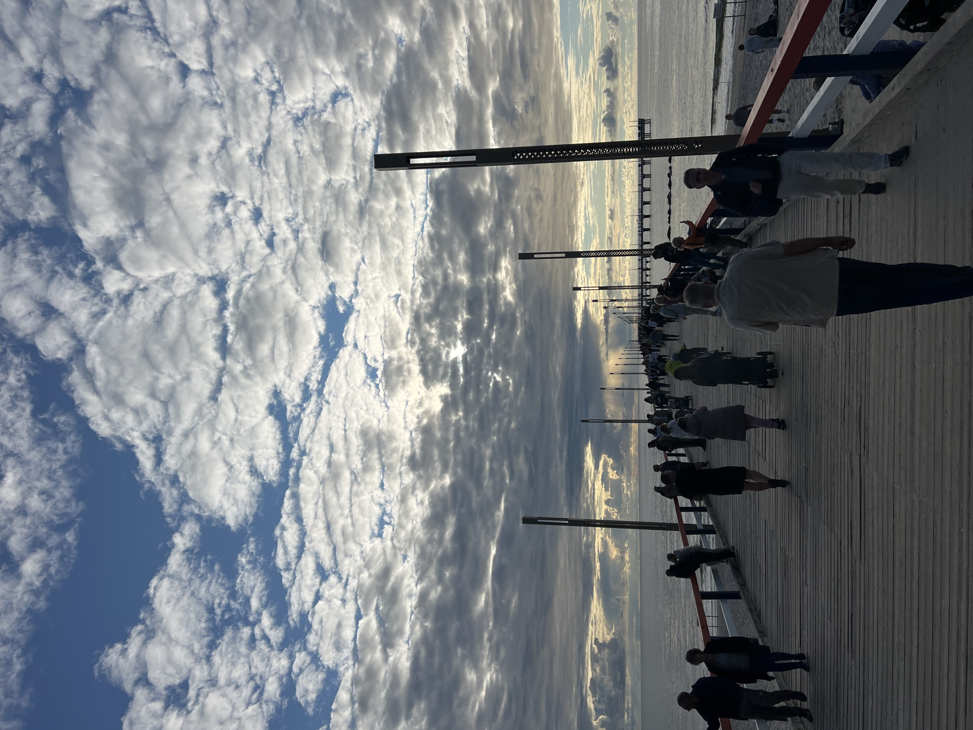 The image captures a scene on the Palanga Pier in Palanga, Lithuania. The pier, constructed of weathered wooden planks, stretches into the distance towards the Baltic Sea, which is visible on the right. A railing with red and white elements lines the side of the pier. A significant number of people are walking in both directions along the pier, silhouetted against the bright sky and sea. The sky is filled with dramatic, fluffy clouds, some of which are backlit by the setting or rising sun, creating shafts of light. Tall, modern lampposts with decorative perforated sides are positioned at intervals along the pier. The overall atmosphere suggests a late afternoon or early evening promenade.