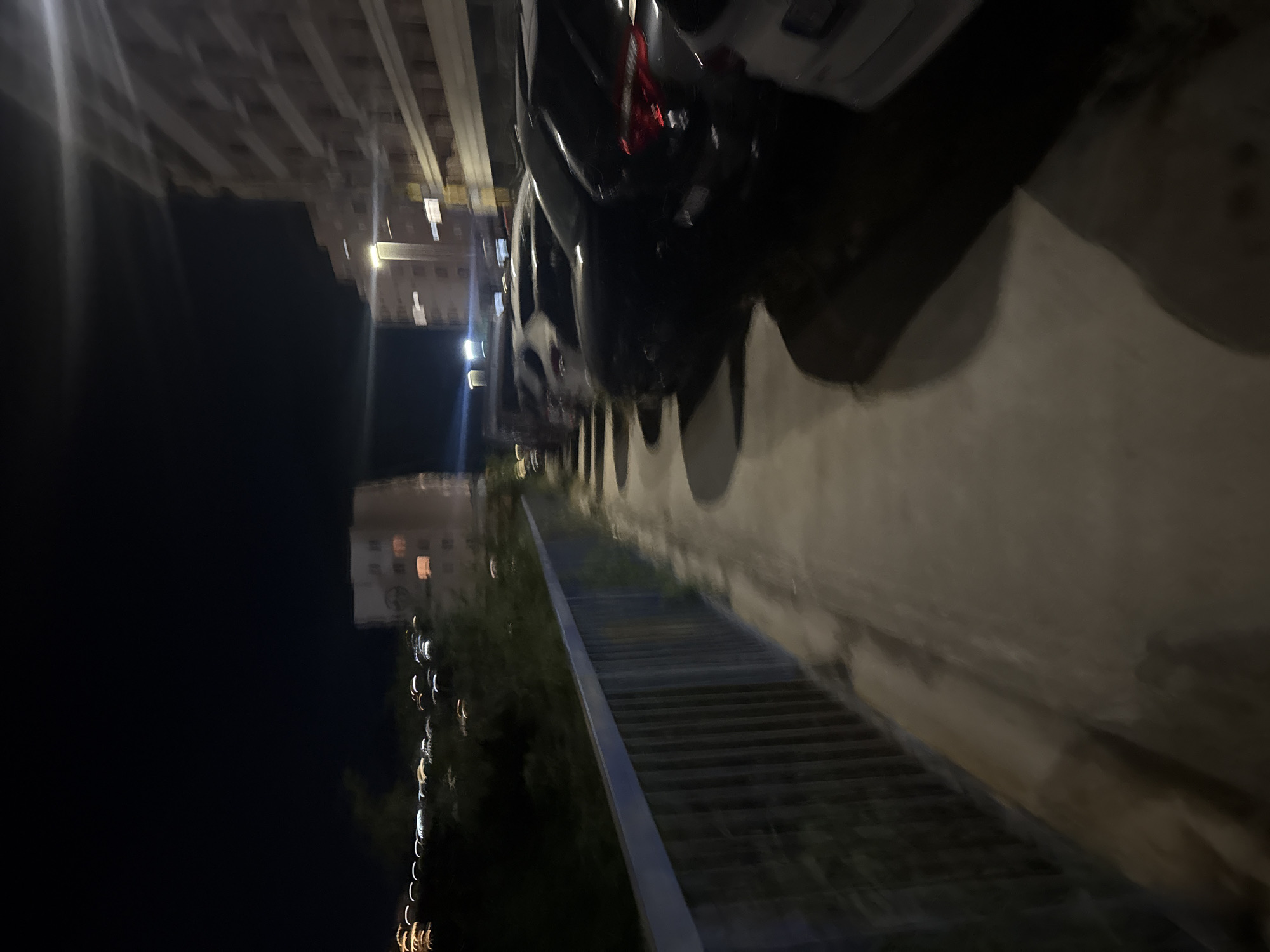 The image shows a nighttime view of a street or sidewalk in Boreti, Montenegro. A metal railing runs along the left side of the frame, bordering a pathway. Beyond the railing, there is dark foliage and distant lights from buildings. To the right of the pathway, several cars are parked in a line. The buildings in the background appear to be multi-story residential structures, with some windows illuminated. Streetlights are also visible, casting a glow on the scene. The overall impression is of a residential area at night, likely with some urban development.