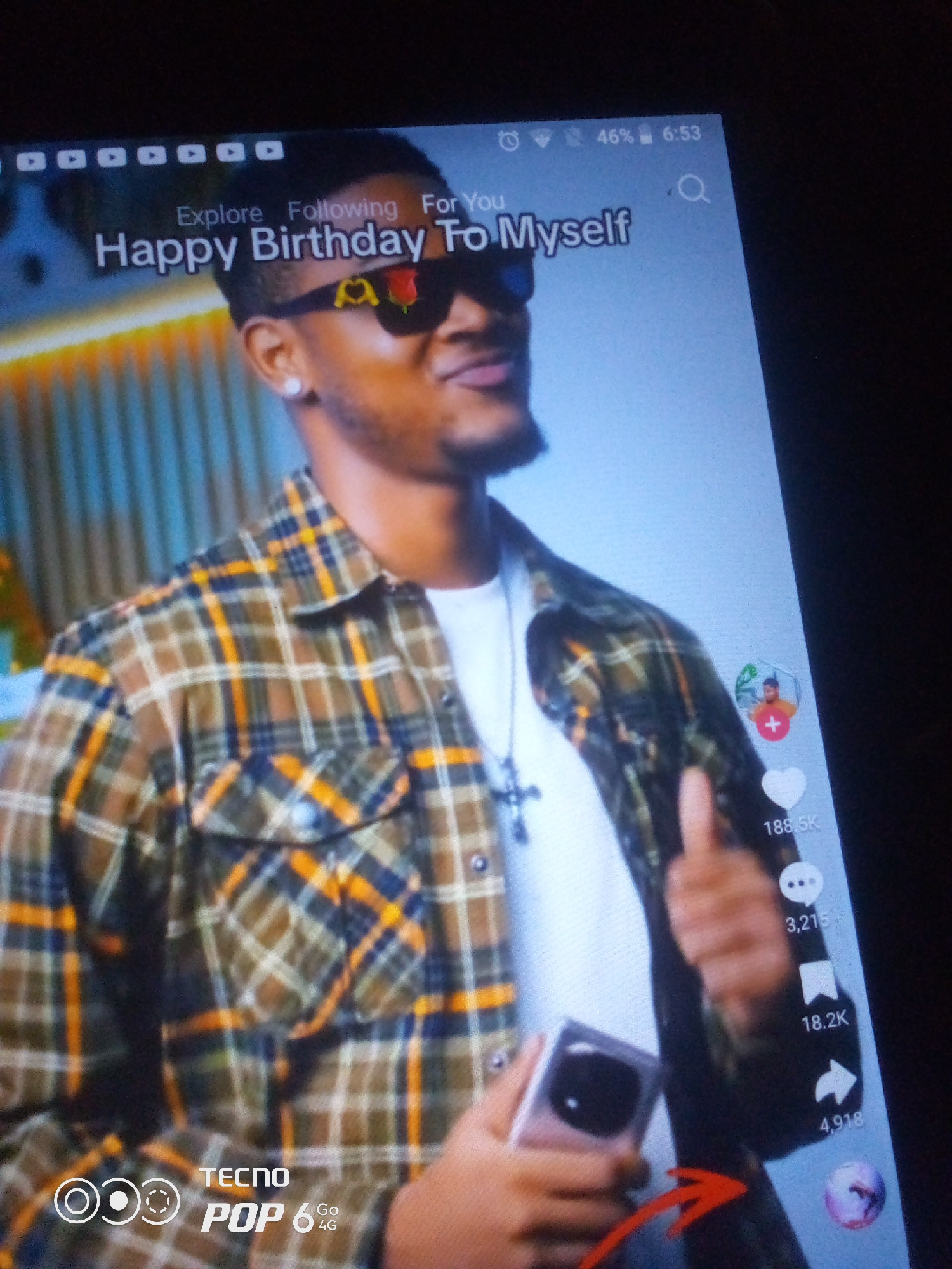 The image shows a close-up of a person wearing sunglasses and a plaid shirt, with a phone or tablet screen in the foreground. The screen displays a video with text that reads "Happy Birthday To Myself". The person's face is partially visible, and they are looking towards the right. There are social media interaction icons on the right side of the screen, including a heart, a comment icon, a bookmark icon, and a share icon, with accompanying numbers indicating engagement. The background of the video appears to be an indoor setting with vertical blinds. The phone itself has a watermark at the bottom left that reads "TECNO POP 6 Go 4G". There is a red arrow pointing from the bottom right of the screen upwards towards the person's hand. The overall image quality suggests it was taken with another camera, possibly a phone, pointed at the screen. The question about the location in Howayi, Nigeria is not supported by any visual information in the image.