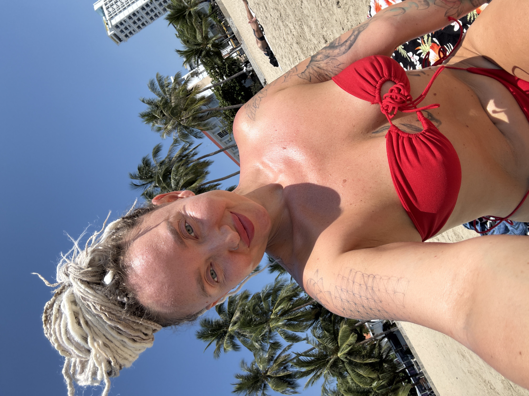 In a bright, sunny outdoor scene on the beach of Nha Trang Ward, Vietnam, a woman with blonde dreadlocks styled in a high bun takes a selfie. She is wearing a red bikini top with a tie detail in the center. Her left arm, visible in the foreground, displays tattoos. In the background, palm trees and white buildings are visible, along with another person lying on a towel on the sand. The sky is clear and blue.