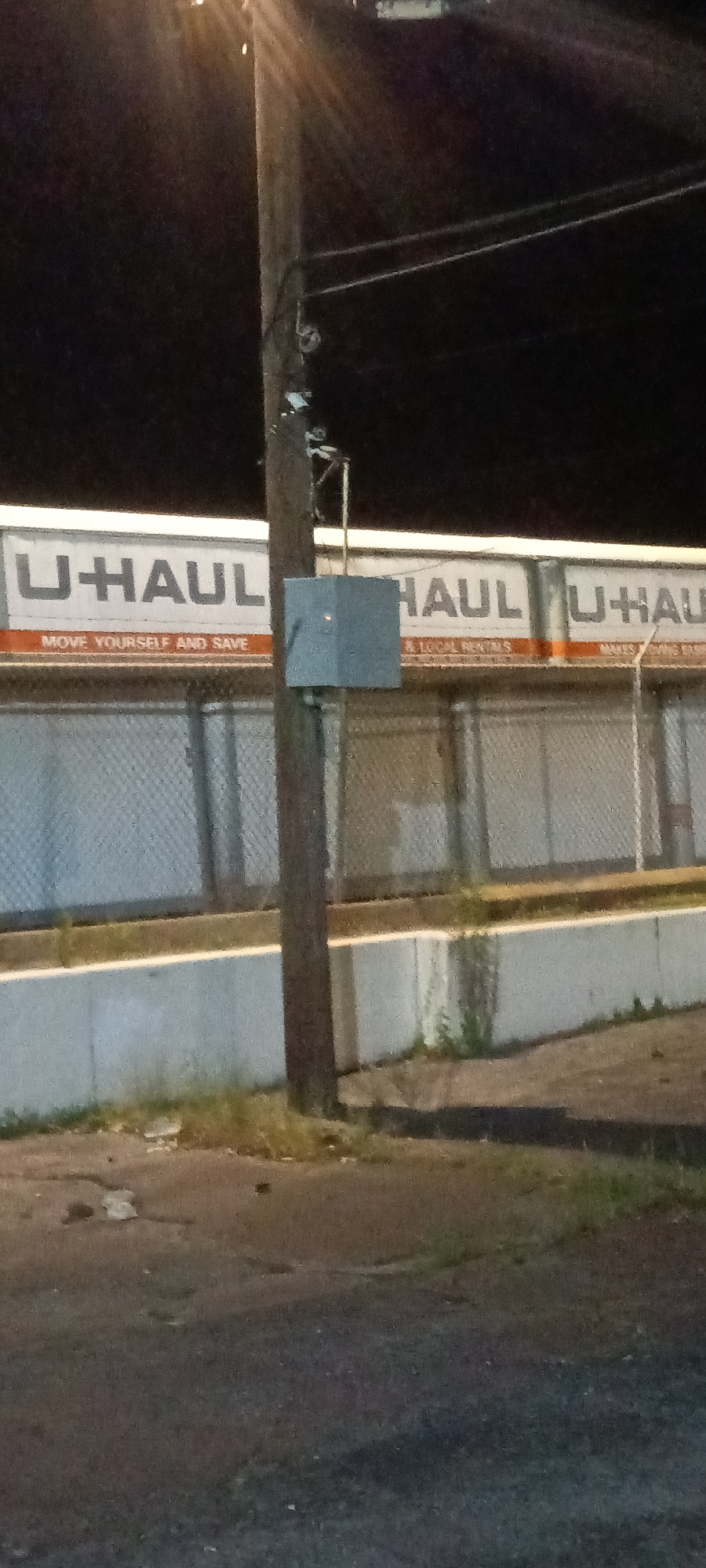 The image depicts a U-Haul facility at night in Fort Smith, Arkansas, United States. The facility is identifiable by the large "U-HAUL" signage on the building. A wooden utility pole is prominently positioned in the foreground, featuring a square metal box attached to it. Below the U-Haul signage is a concrete retaining wall, and there is a paved surface with some vegetation visible. The overall lighting is dim, suggesting it is late evening or night.