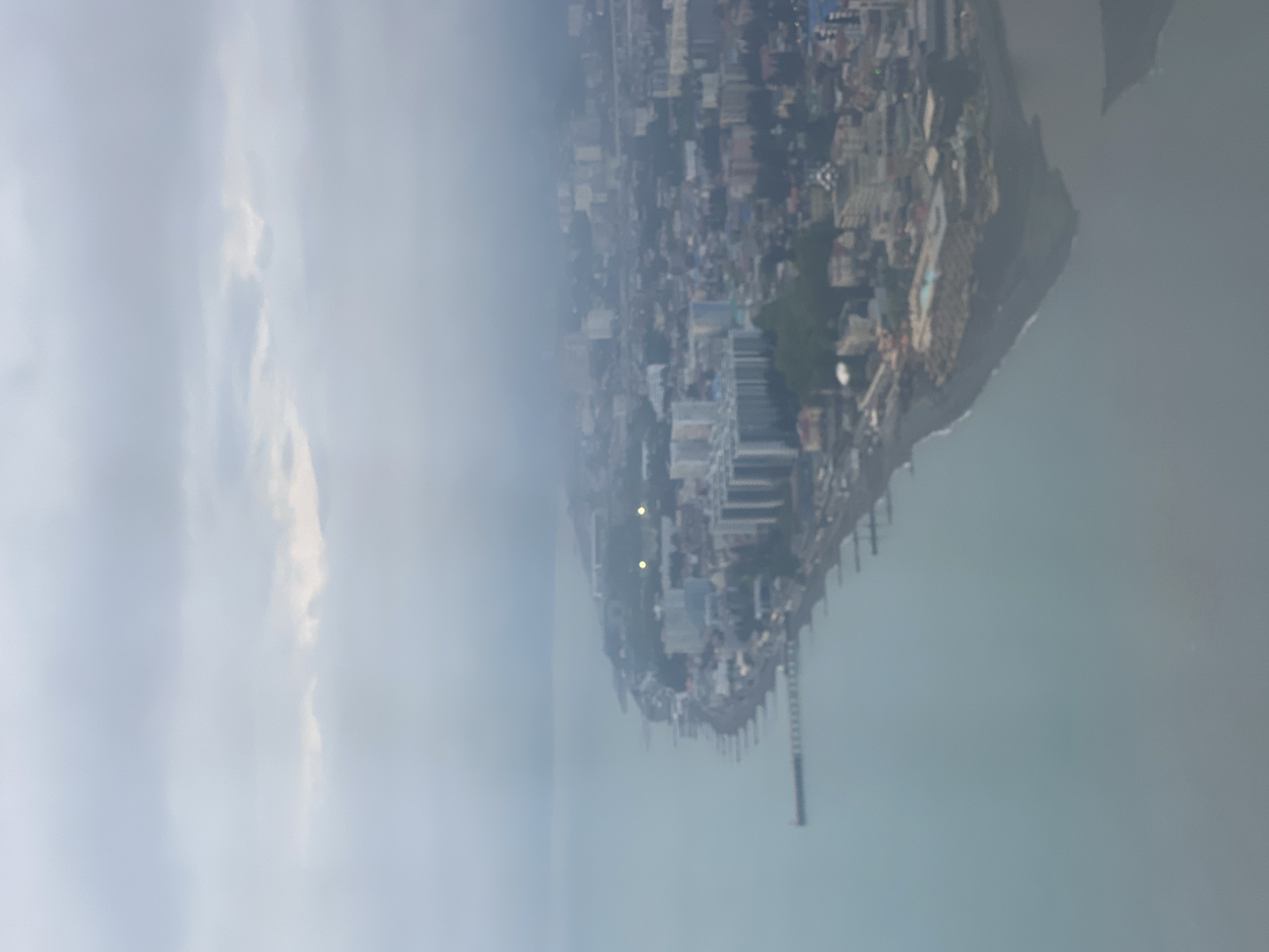 An aerial shot shows a city on a peninsula with a pier extending into the water. The city is densely packed with buildings, and a hazy atmosphere obscures some details. The sky is overcast, and the water is calm. Sochi, Russia.