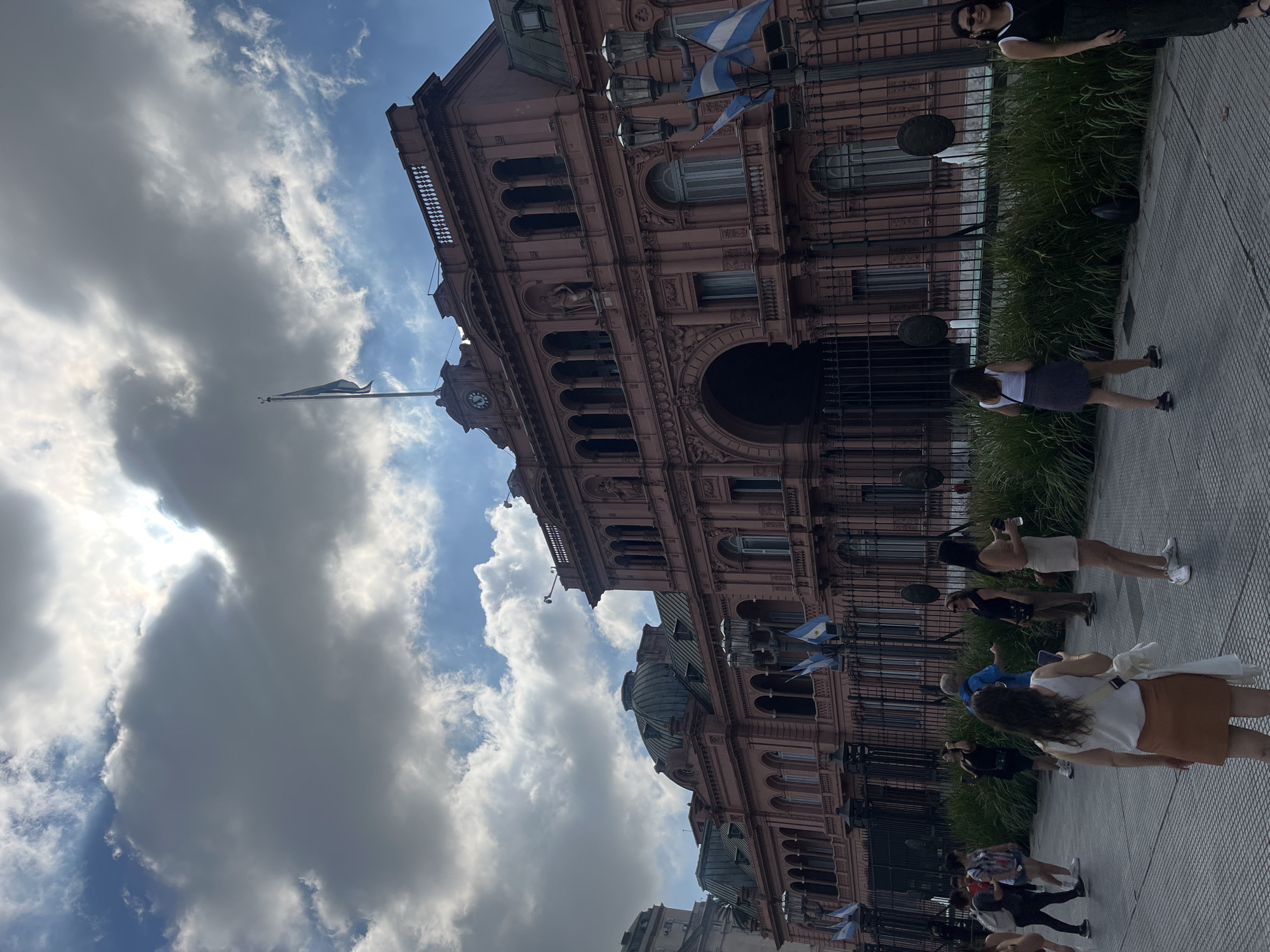 The image captures the Casa Rosada in Buenos Aires, Argentina, from a ground-level perspective. The pink-colored building occupies a significant portion of the frame, showcasing its architectural details such as arches, windows, balconies, and decorative elements. The Argentine flag flies atop a flagpole, with multiple smaller flags adorning the building's facade. A wrought iron fence separates the building from the public sidewalk.
A group of pedestrians occupies the sidewalk, with some standing and others walking. They appear to be tourists or visitors, as some are seen holding phones, potentially taking pictures of the building. Lush green bushes are planted behind the fence. The sky is partially cloudy, with patches of blue visible between the clouds. The lighting is bright and appears to be daytime. The angle of the building and sky suggests the image was taken from a slightly tilted position.
