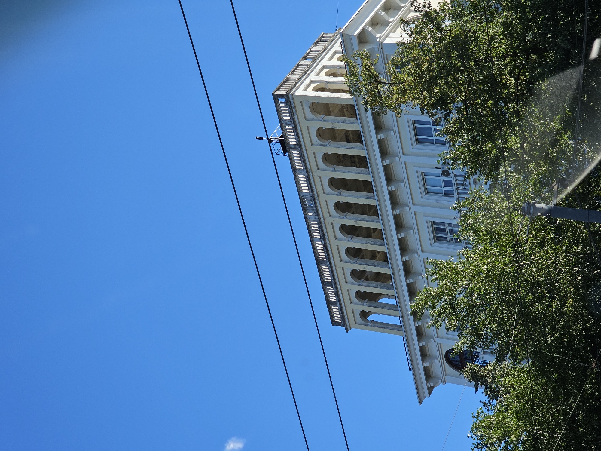 The image shows a building with a colonnaded upper story against a clear blue sky. The building appears to be pale yellow and has white architectural details. There are several horizontal power lines crossing the sky in front of the building. Lush green trees partially obscure the lower floors of the building. The top of the building features a railing with vertical supports and a small structure that resembles an antenna or weather vane. The architectural style suggests it could be located in Nizhny Novgorod, Russia.