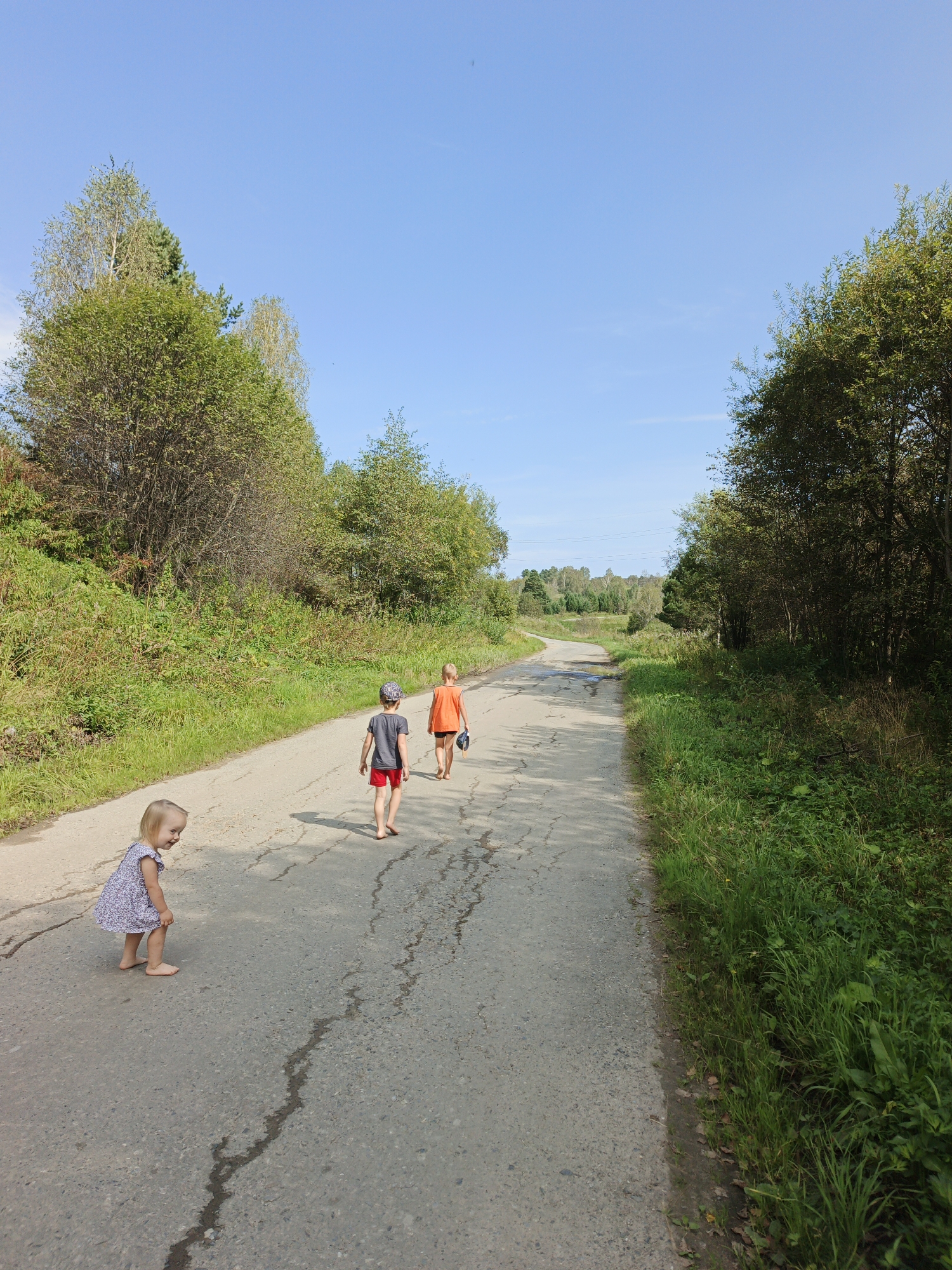 Three children are walking on a paved road in a rural area. The road is lined with green grass and trees. The sky is blue and clear. The child in the foreground is a young girl in a white dress. She is looking back over her shoulder. The two children behind her are boys. One is wearing a grey t-shirt and red shorts, and the other is wearing an orange t-shirt and black shorts. They are walking away from the camera. The location is Urtiushka, Russia.