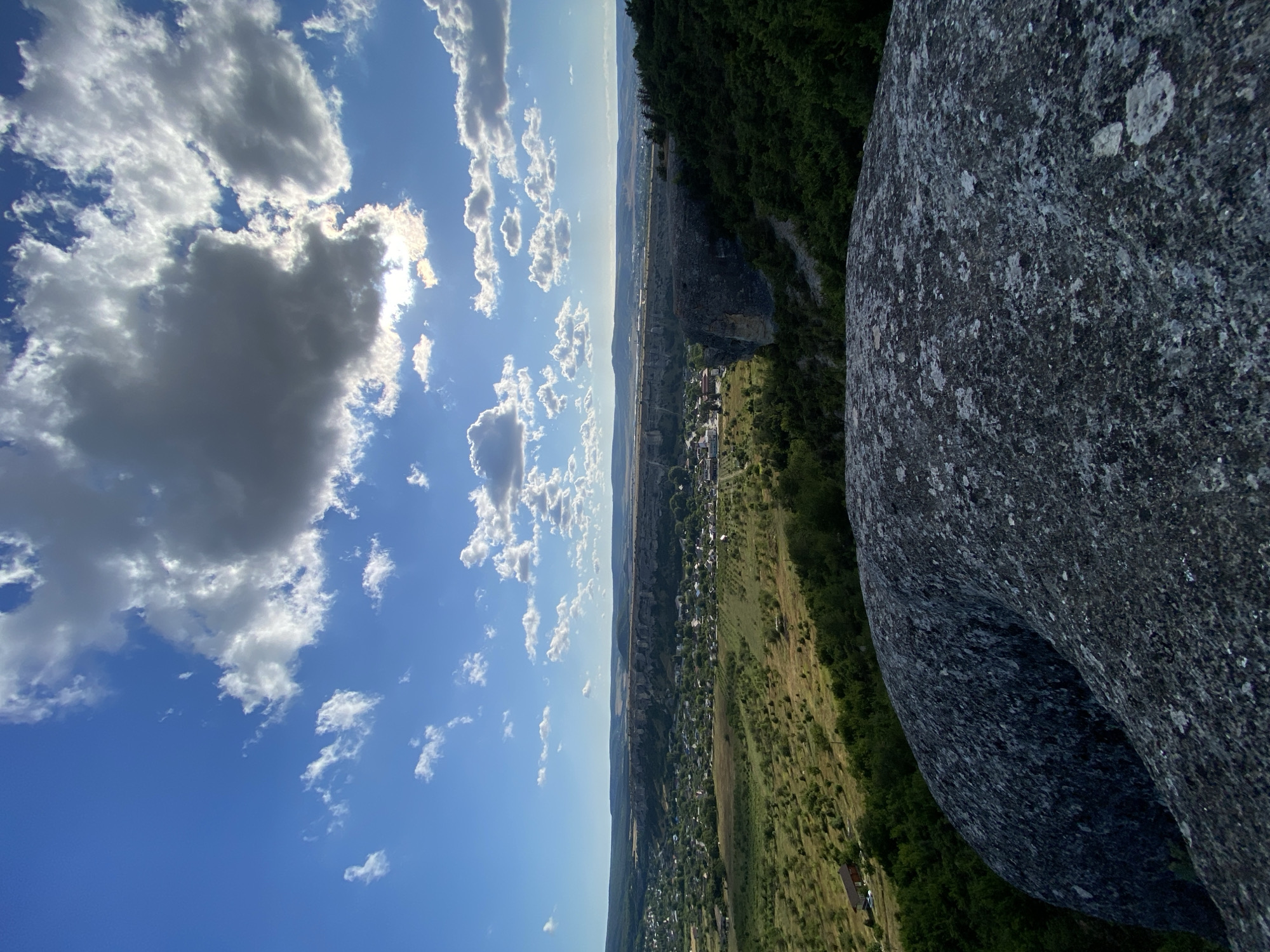 The image presents a panoramic view from a high vantage point, likely a rocky outcrop in the vicinity of Krasnomakskoye, Ukraine. In the foreground, a large, textured gray boulder dominates the lower portion of the frame, its surface rough and uneven. Beyond this boulder, the landscape unfolds. A green, rolling valley stretches into the distance, dotted with scattered trees and houses, indicative of a rural or semi-rural settlement. Further back, a prominent cliff face runs horizontally across the middle ground, with a village nestled at its base. The village appears to consist of numerous buildings with red-tiled roofs. The background features a hazy horizon with undulating hills and a broad expanse of sky filled with a mix of white, puffy cumulus clouds and darker, more substantial clouds, some of which are backlit by the sun. The overall impression is one of natural beauty and tranquility, with a sense of elevation and expansive views. There are no people or animals visible in the image.