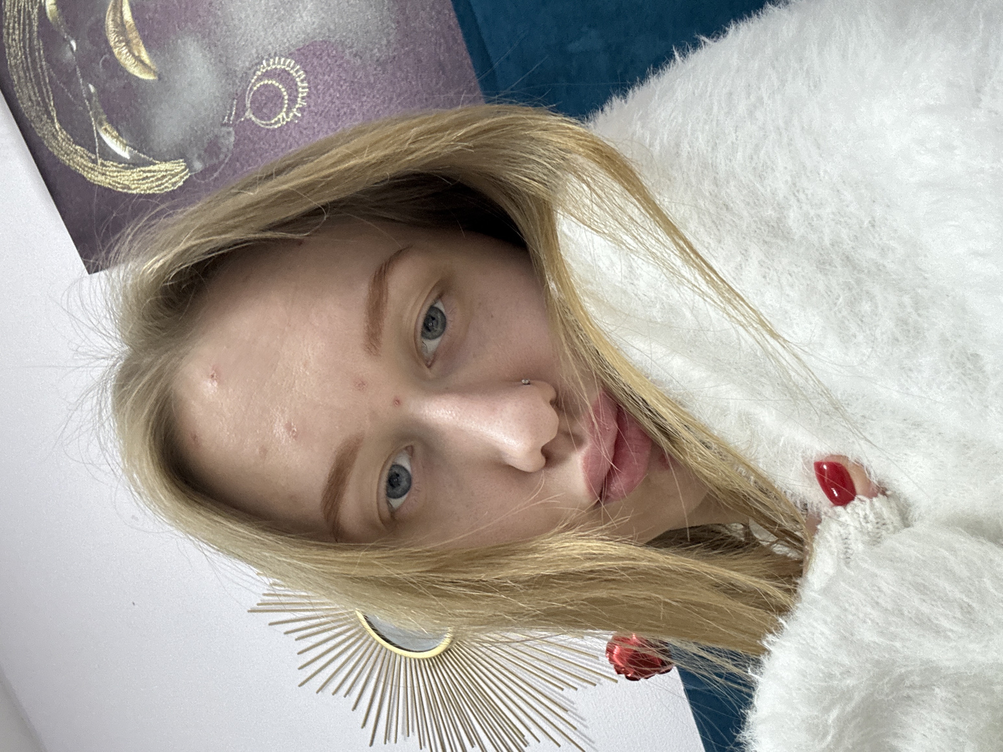 A selfie of a light-skinned person with blonde hair, wearing a white fuzzy sweater. The person has blue eyes, a nose piercing, and red nail polish on at least one visible fingernail. The background is a mix of items, including a purple abstract art piece, a teal-colored object, and a gold sunburst mirror. No information is available to determine the location as Rostov-on-Don, Russia.