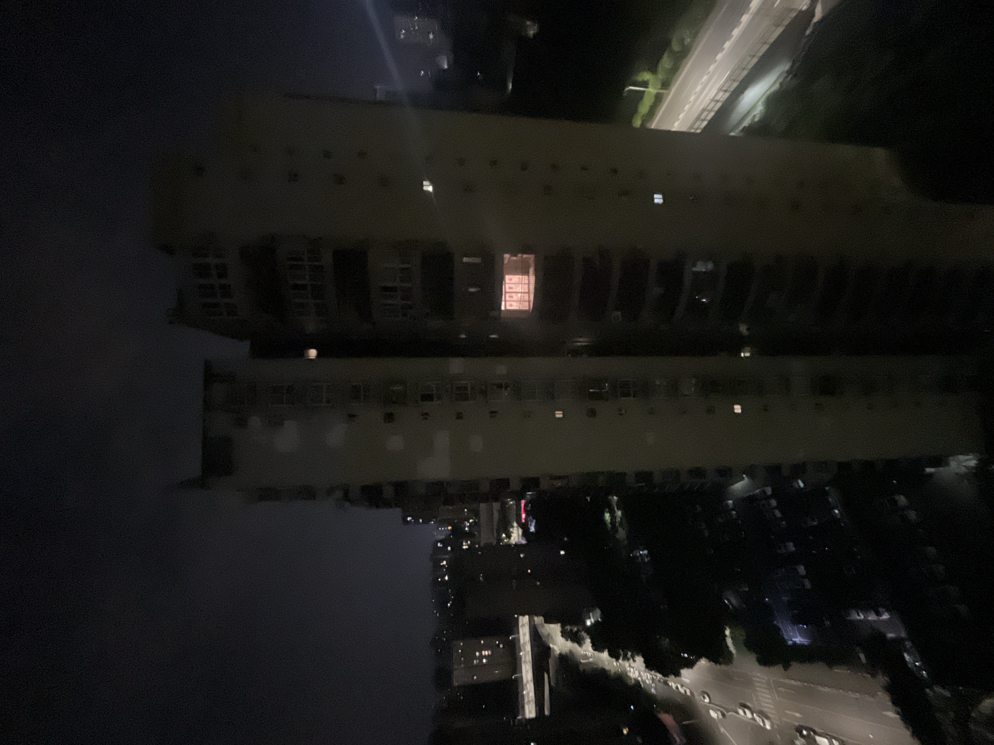 This nighttime image captures an urban scene in Jiulongpo District, China, dominated by two tall residential buildings in the midground, seen from an elevated perspective, likely from another building. The sky above is dark and overcast.
The building on the left is partially visible, while the one on the right stands prominently, both featuring multiple stories with visible balconies and numerous windows. A few windows are illuminated, with one particular window on the rightmost building displaying a distinct, warm, reddish-orange glow, suggesting an interior light source.
Below the buildings, a network of roads is visible, illuminated by streetlights and the headlights/taillights of several vehicles, some parked along the street, others moving, appearing as light trails due to the low-light exposure. A raised road or bridge crosses horizontally in the mid-distance on the left side of the frame. Scattered trees and dark foliage line the roads and fill in gaps between structures. In the far background, the faint outlines of other buildings and city lights extend into the distance. No discernible individuals are present in the image.