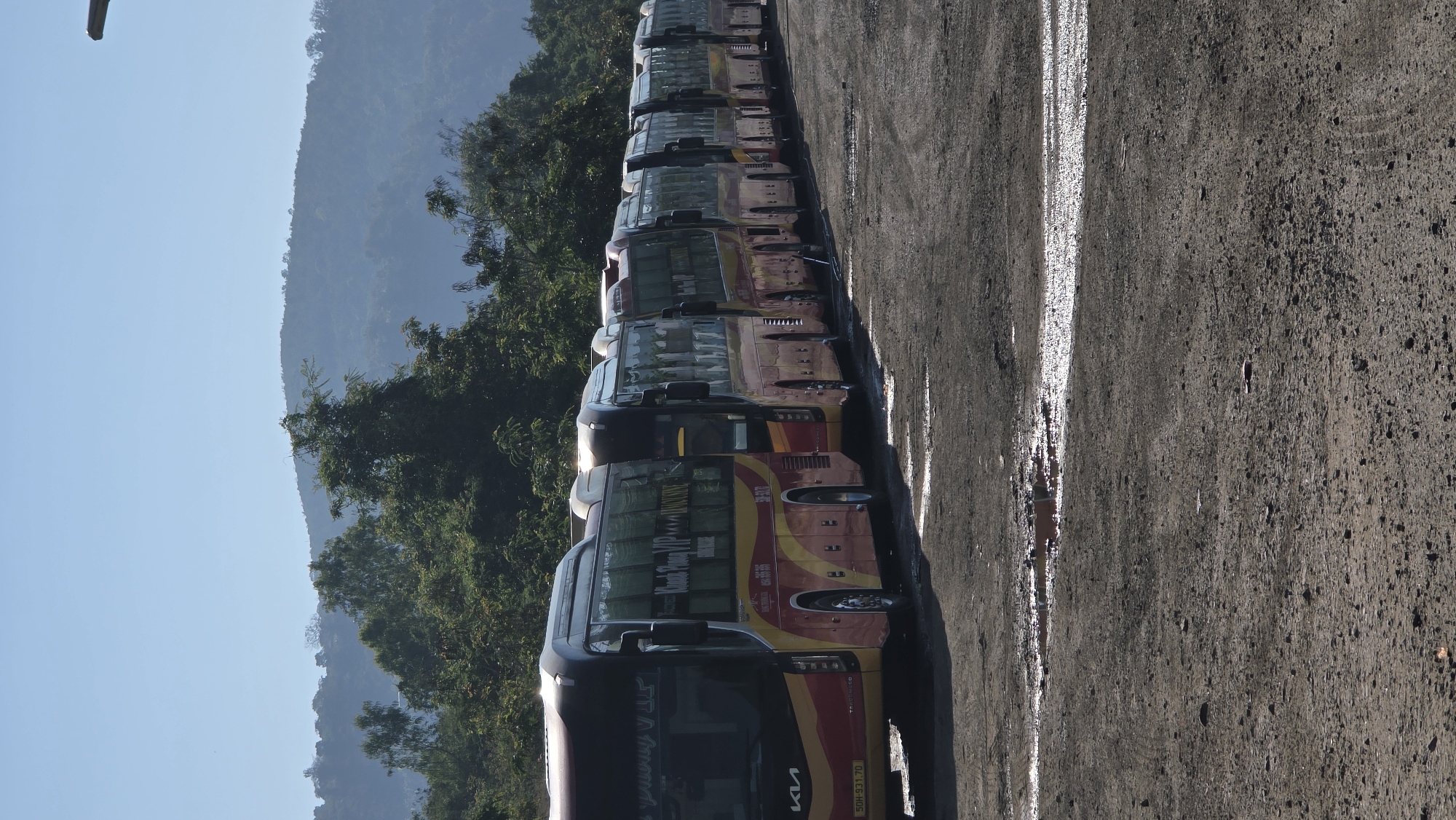 A row of buses is parked in an open lot. The buses are red and yellow with Vietnamese writing. The license plate on the front bus is 50H.931.70. Trees and mountains are in the background, visible behind the buses. The parking lot is gray and cracked. The sky is light blue. This is likely a bus depot in Bắc Nha Trang, Vietnam.