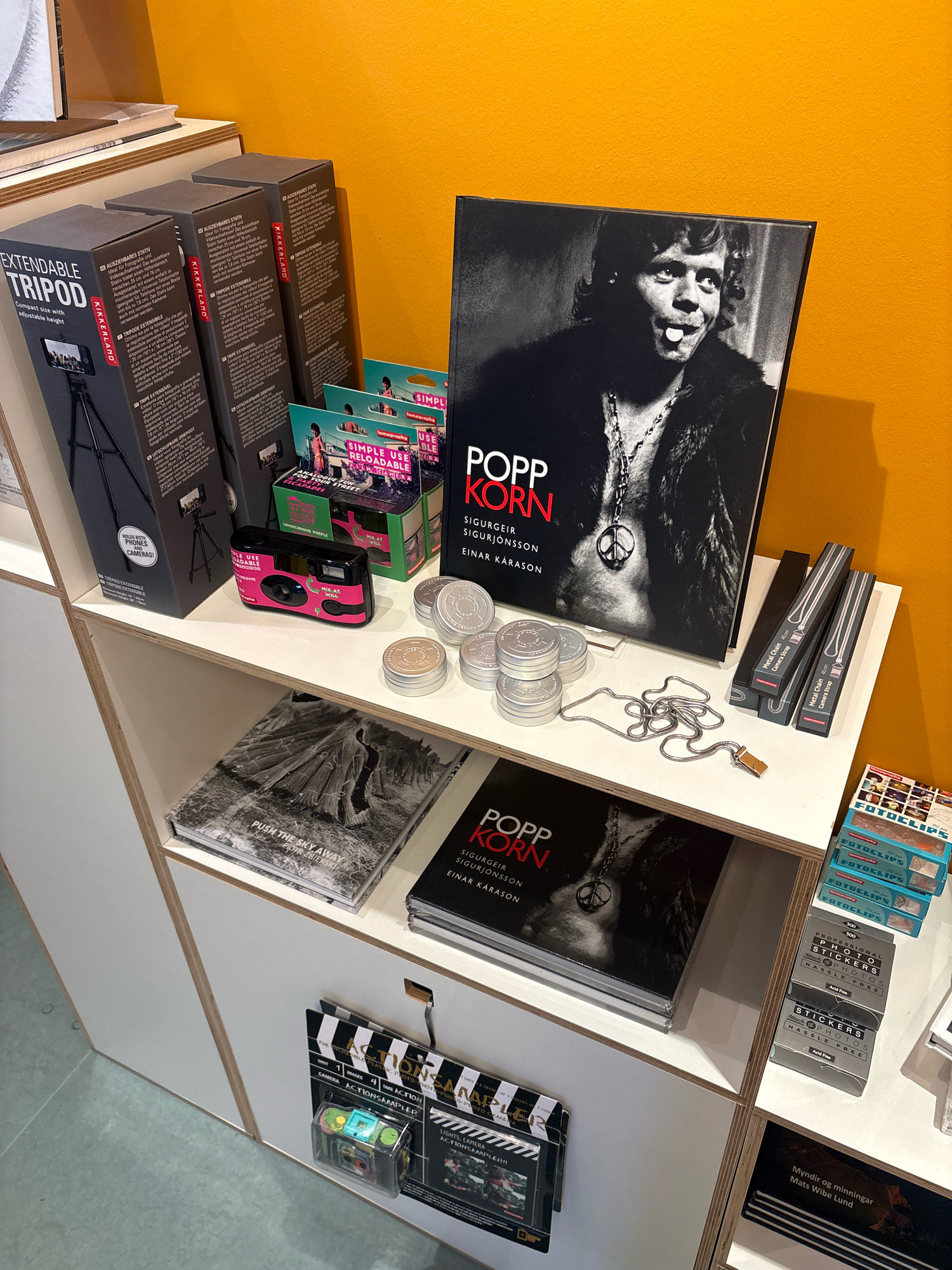 The image displays a retail shelf filled with photography-related merchandise. On the top shelf, three boxed extendable tripods are visible, along with several small boxes labeled "SIMPLE USE RELOADABLE CAMERA" and a large, black hardcover book with the title "POPP KORN SIGURGEIR SIGURJÓNSSON EINAR KARASON". To the right of the book are two boxes of metal camera chains. The middle shelf holds a black and white photo book titled "PUSH THE SKY AWAY PIOTR ZBIERSKI", another copy of "POPP KORN", and several silver circular containers, possibly for film. A silver snake chain with a gold clasp is draped across this shelf. The bottom shelf features two "ACTIONSAMPLER!!" camera kits and a book with Icelandic text, "Myndir og minningar Mats Wibe Lund". A stack of small blue boxes labeled "FOTOCLIPS" is visible on the far right. The overall setting is a brightly lit retail environment with an orange wall in the background. This scene is consistent with a camera shop or a bookstore that sells photography books and accessories. The presence of Icelandic titles on some of the books and the overall aesthetic suggest the location is in Reykjavik, Iceland.