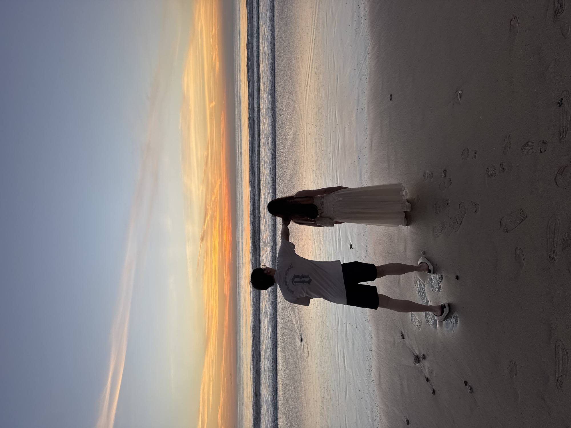 Two individuals are standing on a beach. One person, wearing a white shirt and black shorts, is holding the other person's hand. The second person is wearing a light-colored dress. The sky shows a gradient of colors, from blue to orange, suggesting either sunrise or sunset. The ocean is visible as a dark line in the middle ground. There are footprints in the sand. The location is consistent with a beach in Kuta Selatan, Indonesia.