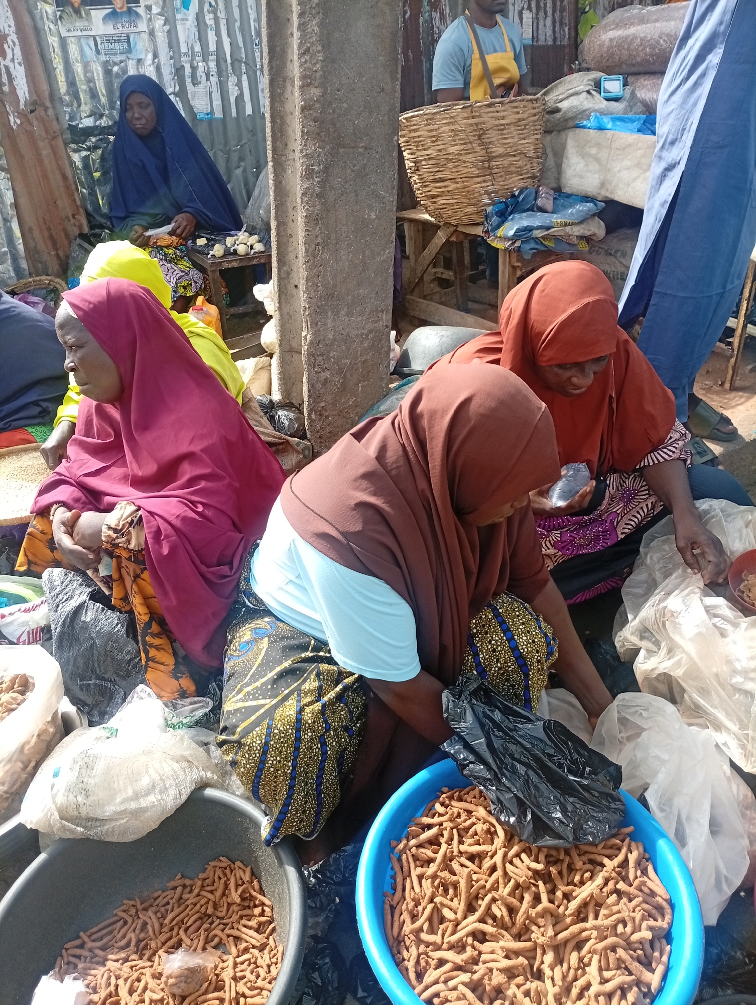 The image captures a scene at a market in Maiburiji, Nigeria. Several women are seated on the ground surrounded by goods for sale. The women wear headscarves in varying colors: brown, red, yellow, and blue. They appear to be sorting or packing items, possibly foodstuff, into plastic bags.
A blue plastic basin filled with brown, elongated, stick-like snacks is prominently in the foreground. Similar items are in plastic bags and another basin to the left. A man in a yellow apron stands in the background. The surroundings suggest a bustling market environment with metal sheeting structures and posters on a nearby wall.